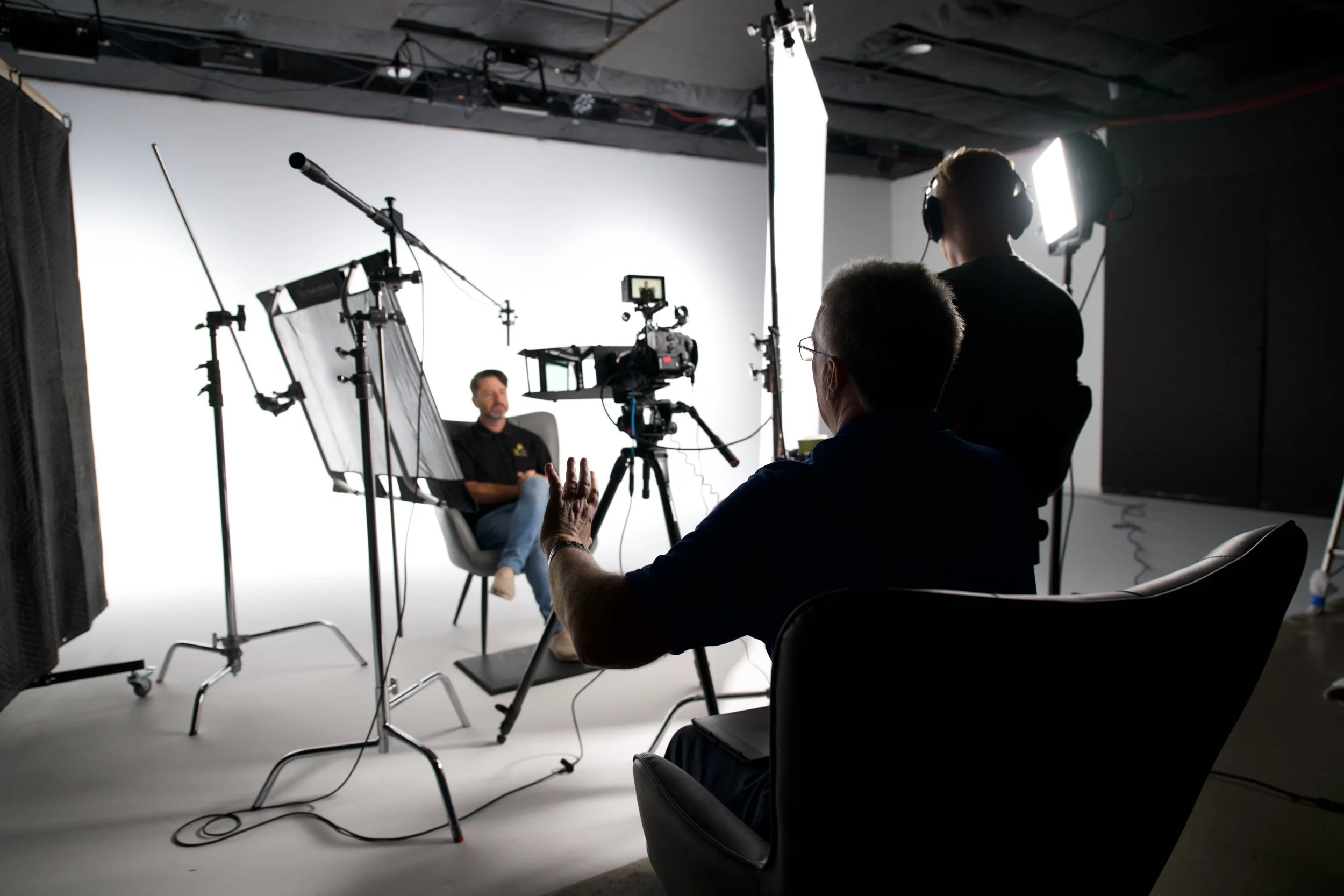 A video interview setup with a man seated in front of a white backdrop, surrounded by camera equipment, lighting, and crew members filming and managing the scene.