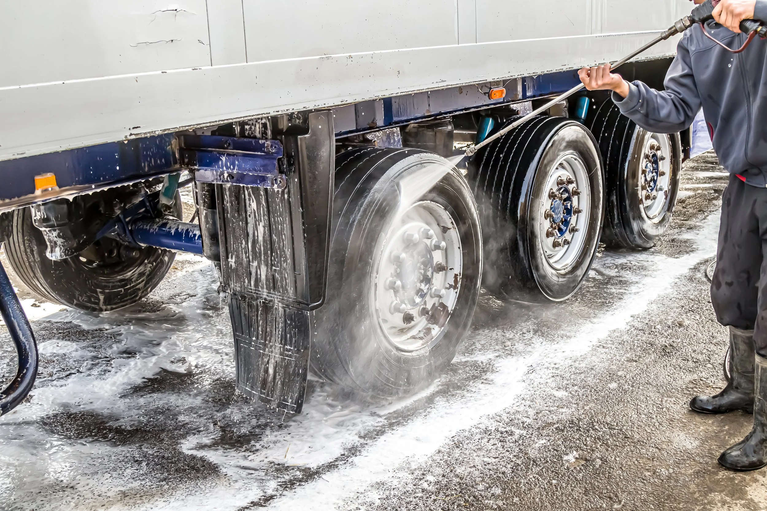 Someone washing the wheels and undercarriage of a large truck with a pressure washer, creating soap suds and water on the ground.