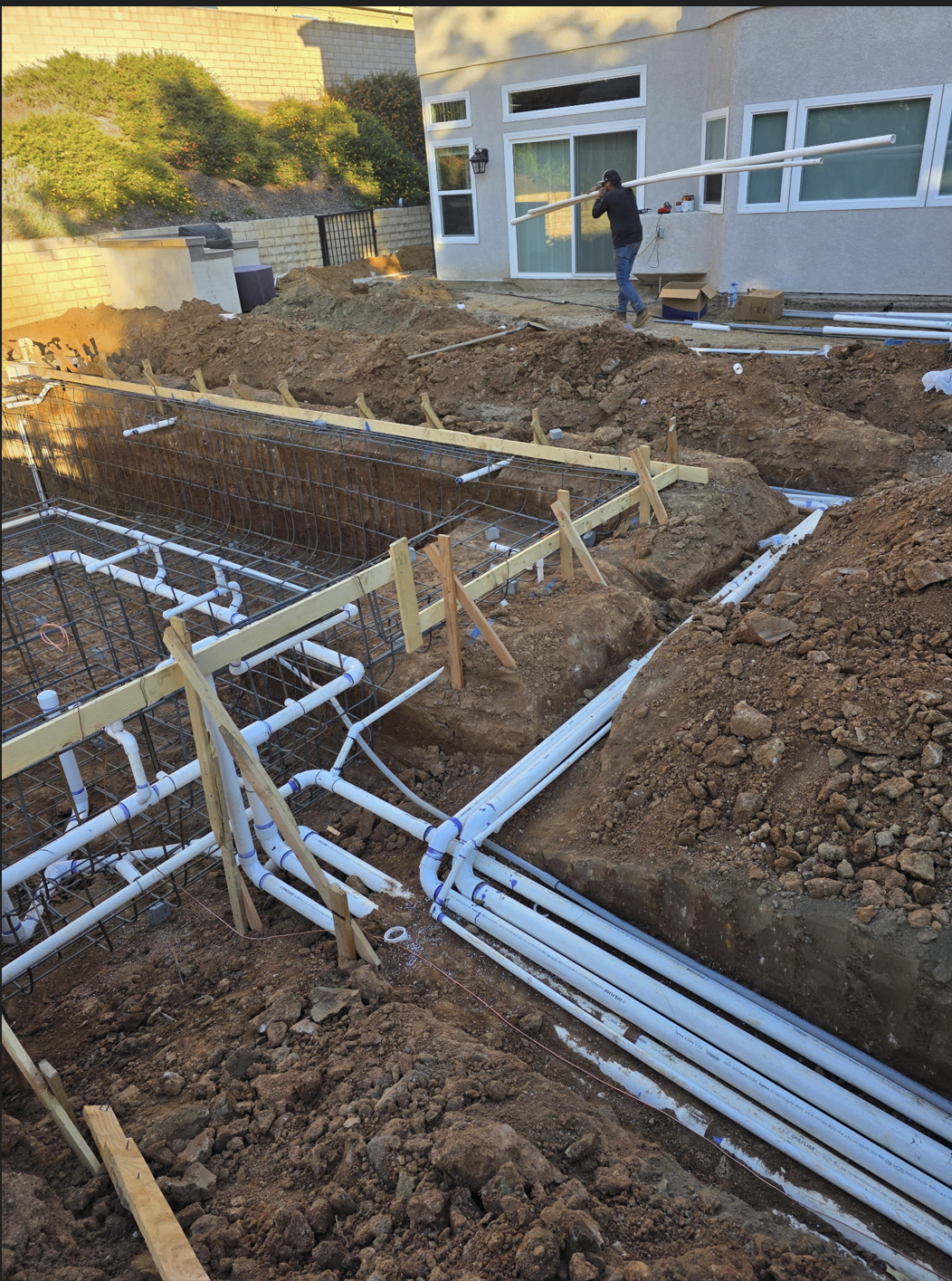 Construction site showing plumbing pipes being installed for a building foundation, with an excavated trench, rebar framework, and worker walking with pipes in hand, near a house with beige exterior walls and windows.