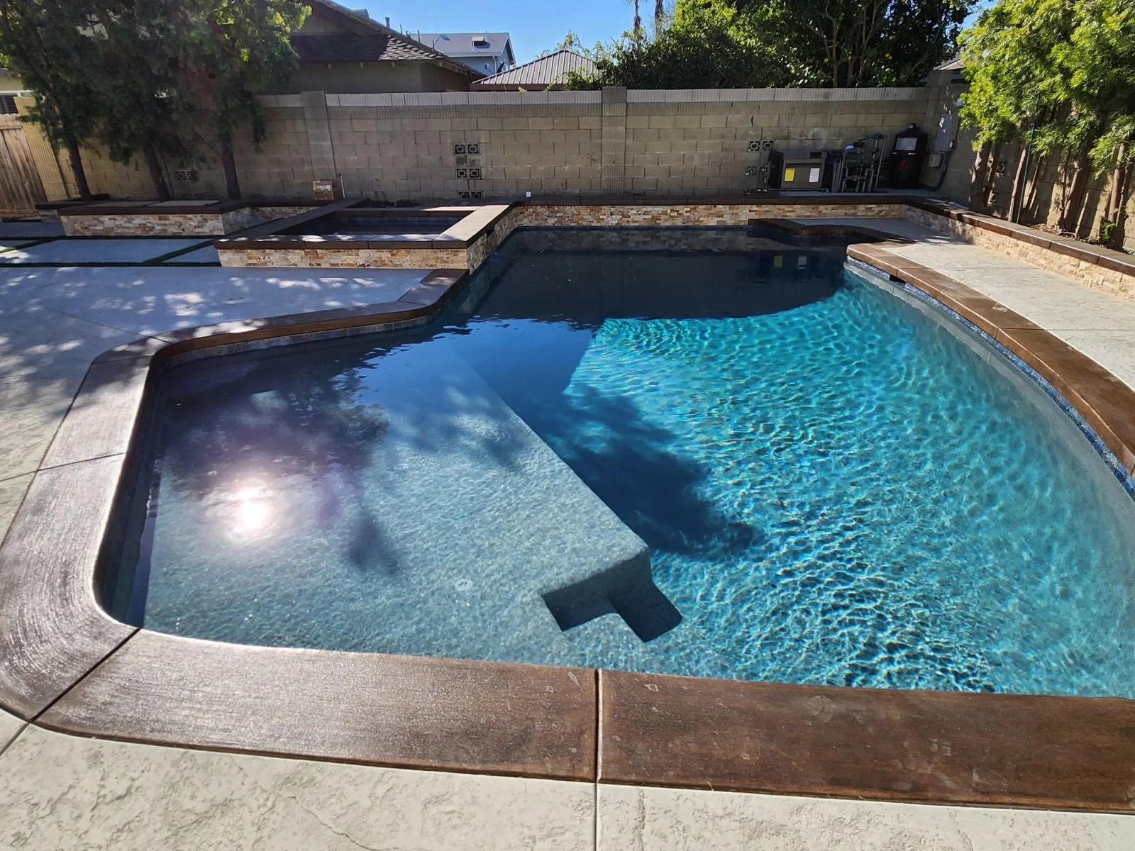 A backyard swimming pool with a built-in hot tub, surrounded by stone and concrete decking, with a brick wall and trees in the background.