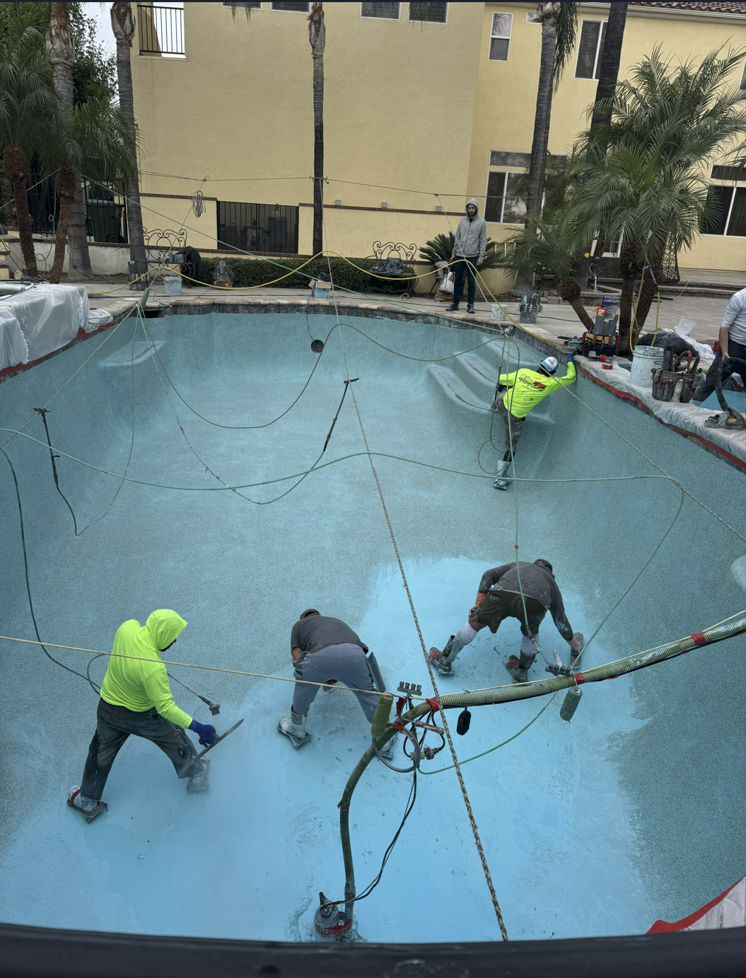 Workers refurbishing a swimming pool with blue surface and surrounding construction equipment, some palm trees and a yellow building in background.
