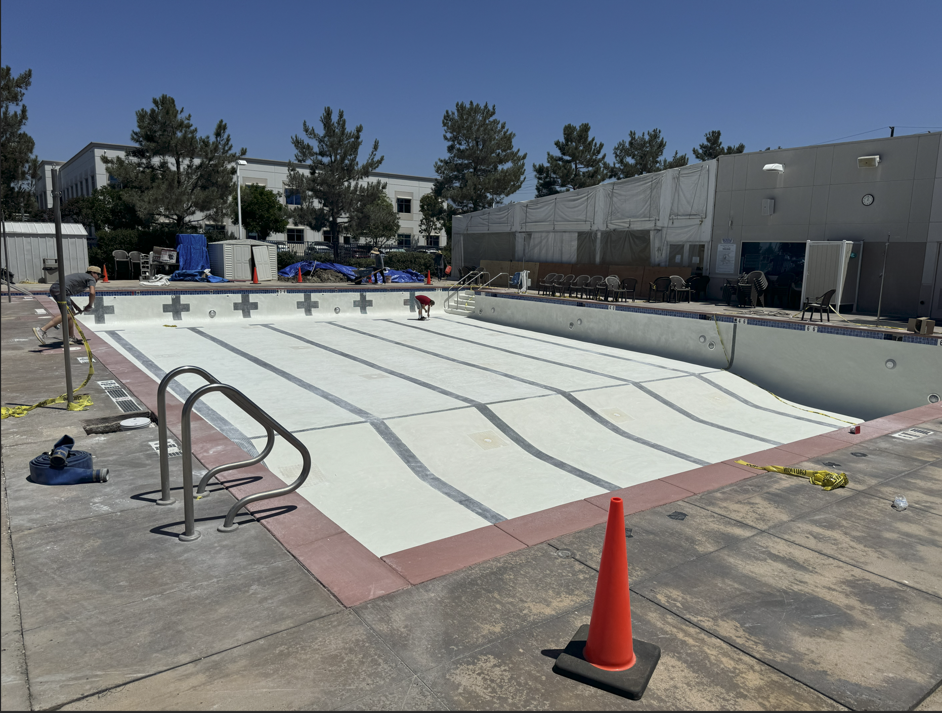 An empty outdoor swimming pool under renovation, with the pool surface being resurfaced. Workers are working on the pool and safety cones are placed around the area, with pool chairs and equipment in the background.