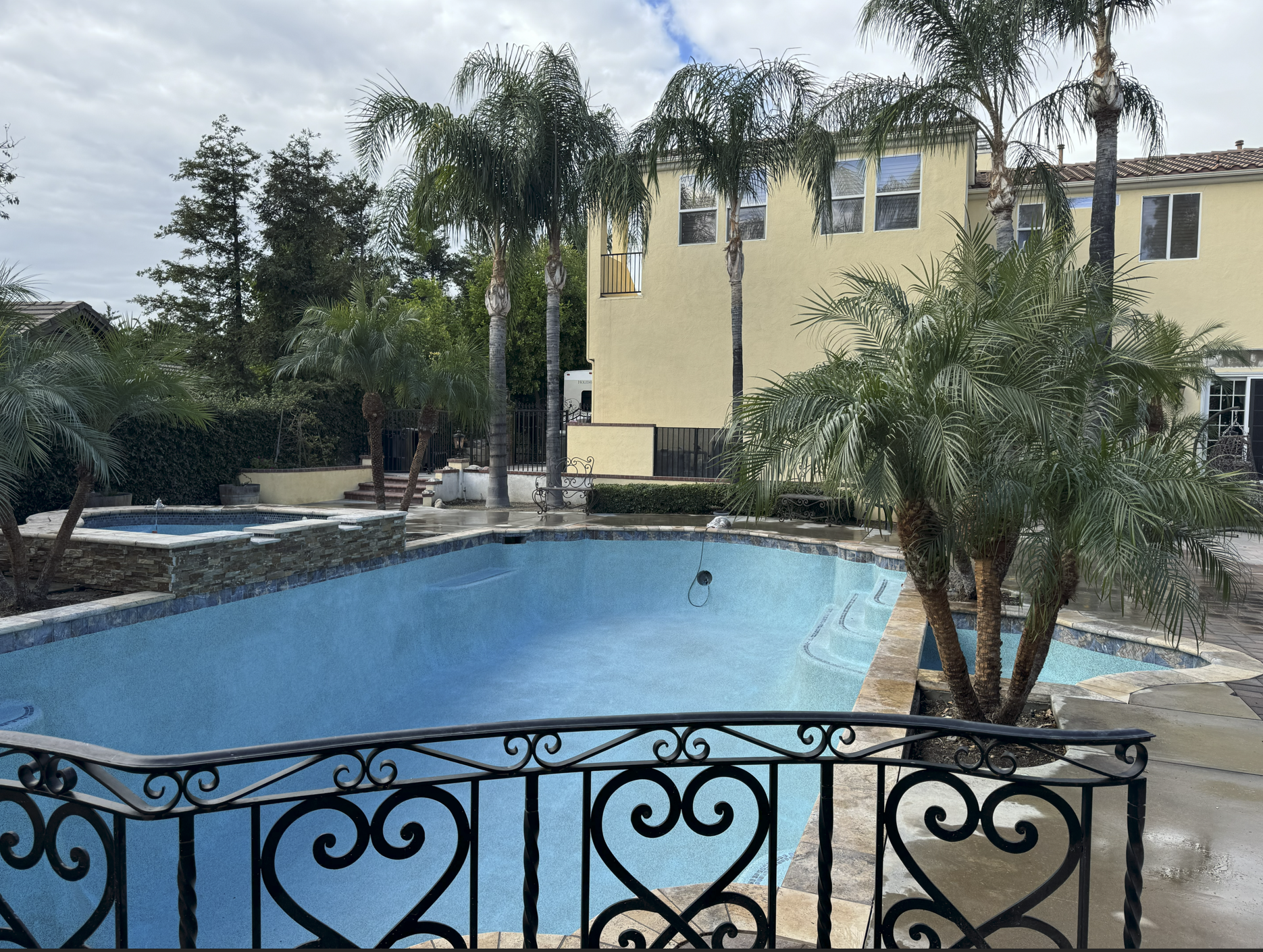 Empty backyard swimming pool with a decorative black metal fence, surrounded by palm trees, a yellow house, and outdoor furniture on a cloudy day.