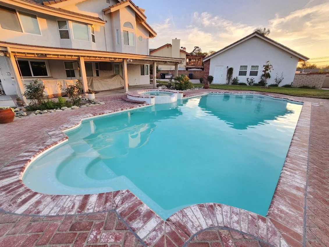 Residential backyard with a kidney-shaped swimming pool, hot tub, brick patio, and lawn, during sunset.