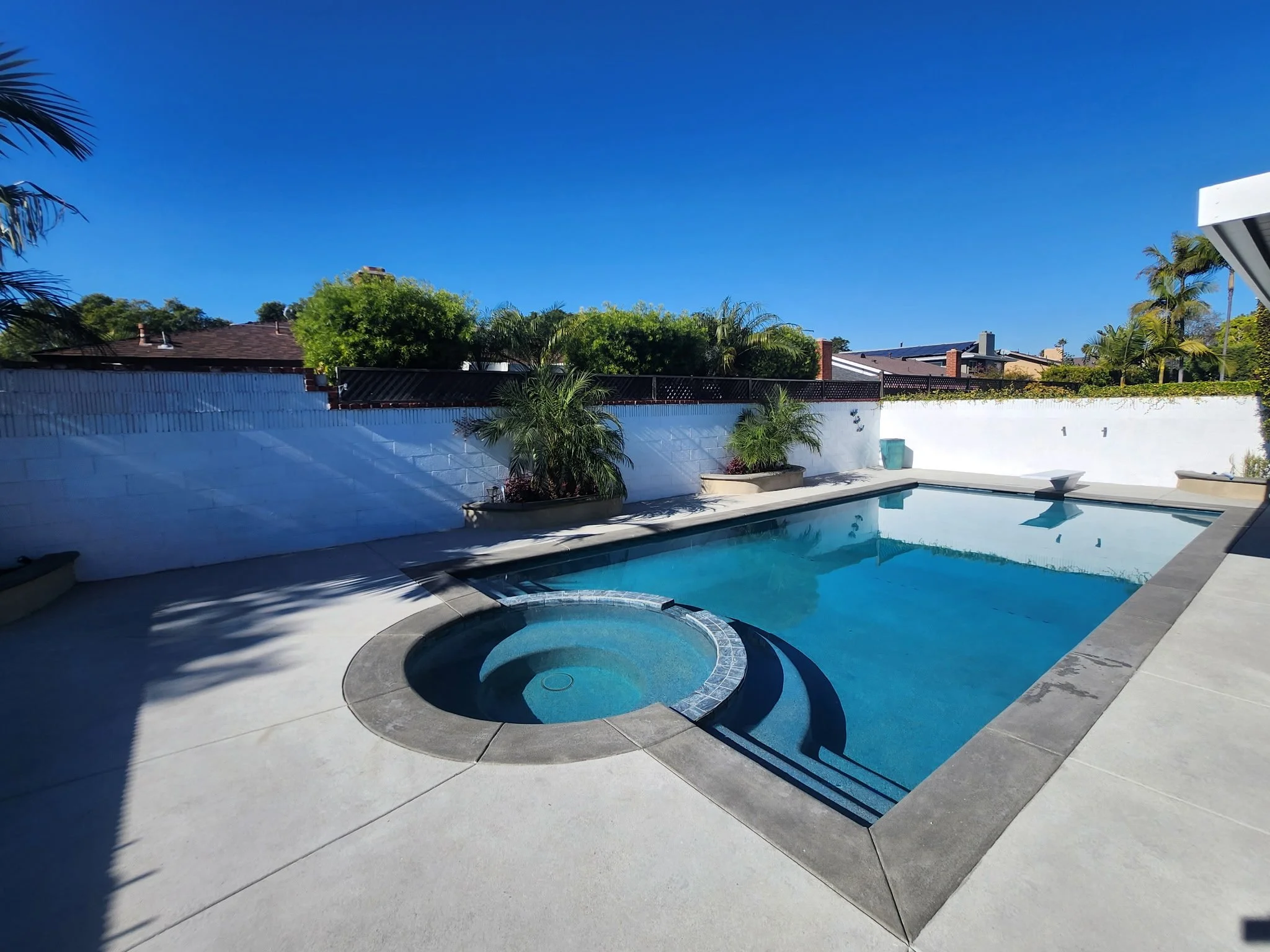Empty backyard swimming pool with a round hot tub in the foreground and a fenced yard with trees in the background under a clear blue sky.