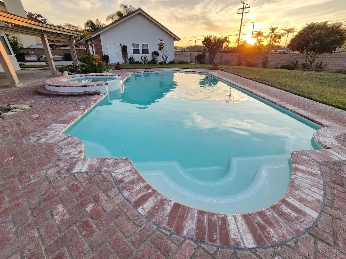 A backyard swimming pool with clear water at sunset, surrounded by brick paving, a grassy lawn, trees, and a white house in the background.