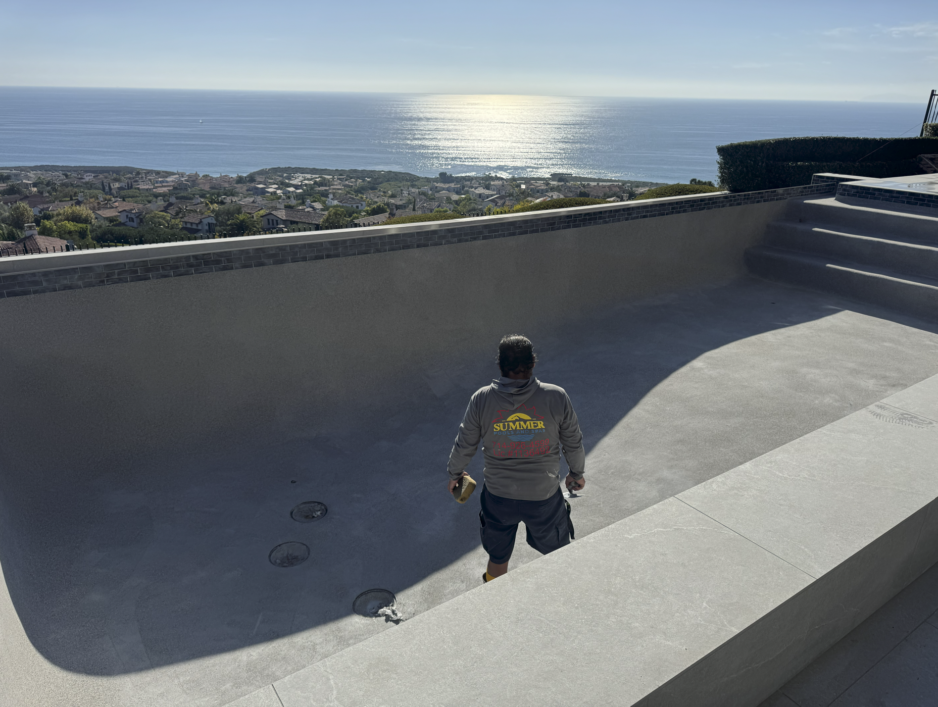 Man standing in a large empty backyard with stairs and a view of the ocean and houses in the distance.