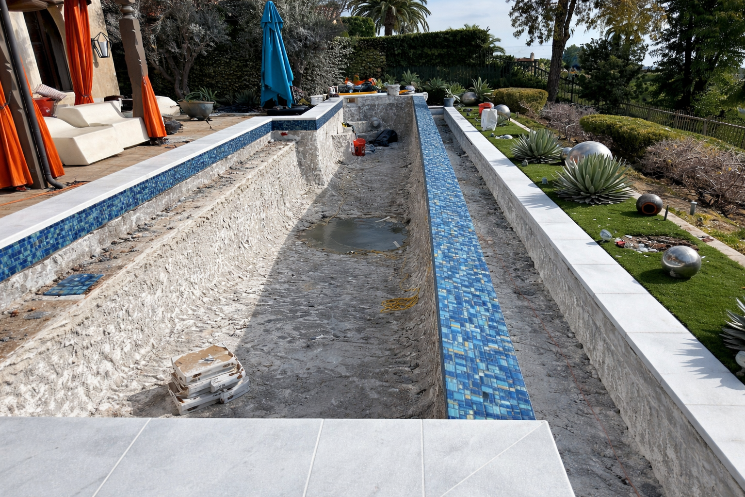 A backyard swimming pool under renovation with tiling in progress, surrounded by a deck with outdoor furniture, umbrellas, and decorative plants, with construction tools and debris present.