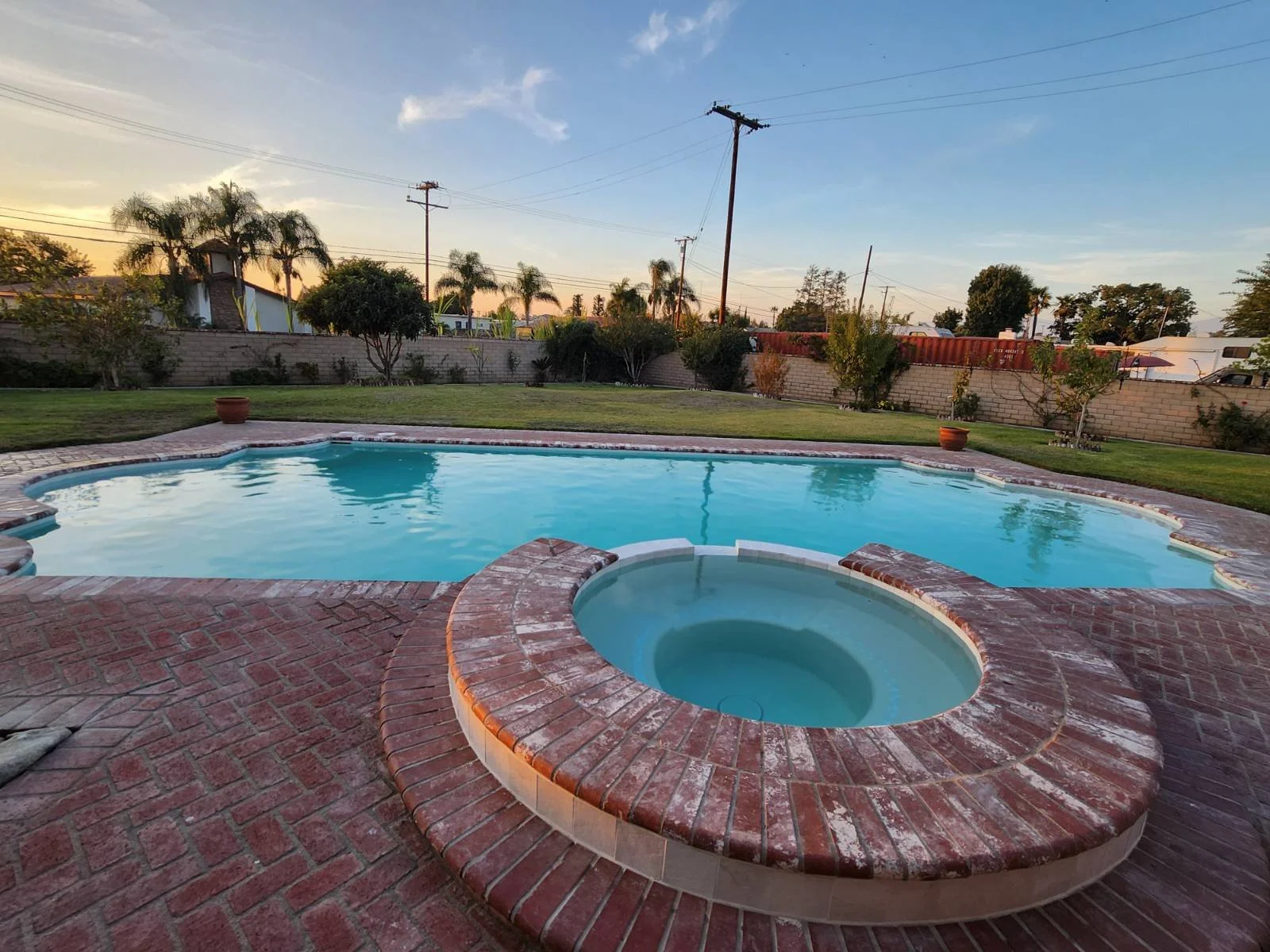 A backyard with a swimming pool and a hot tub, surrounded by a brick patio and a lawn with trees and bushes, under a clear sky at sunset.