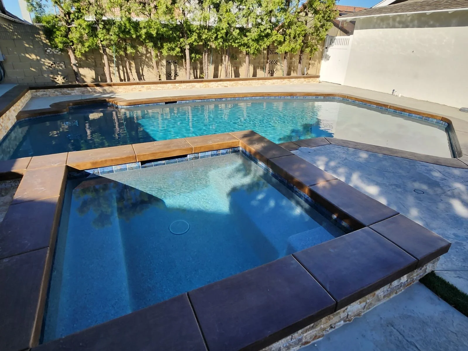 Backyard swimming pool with a hot tub, surrounded by a concrete patio and trees in the background.