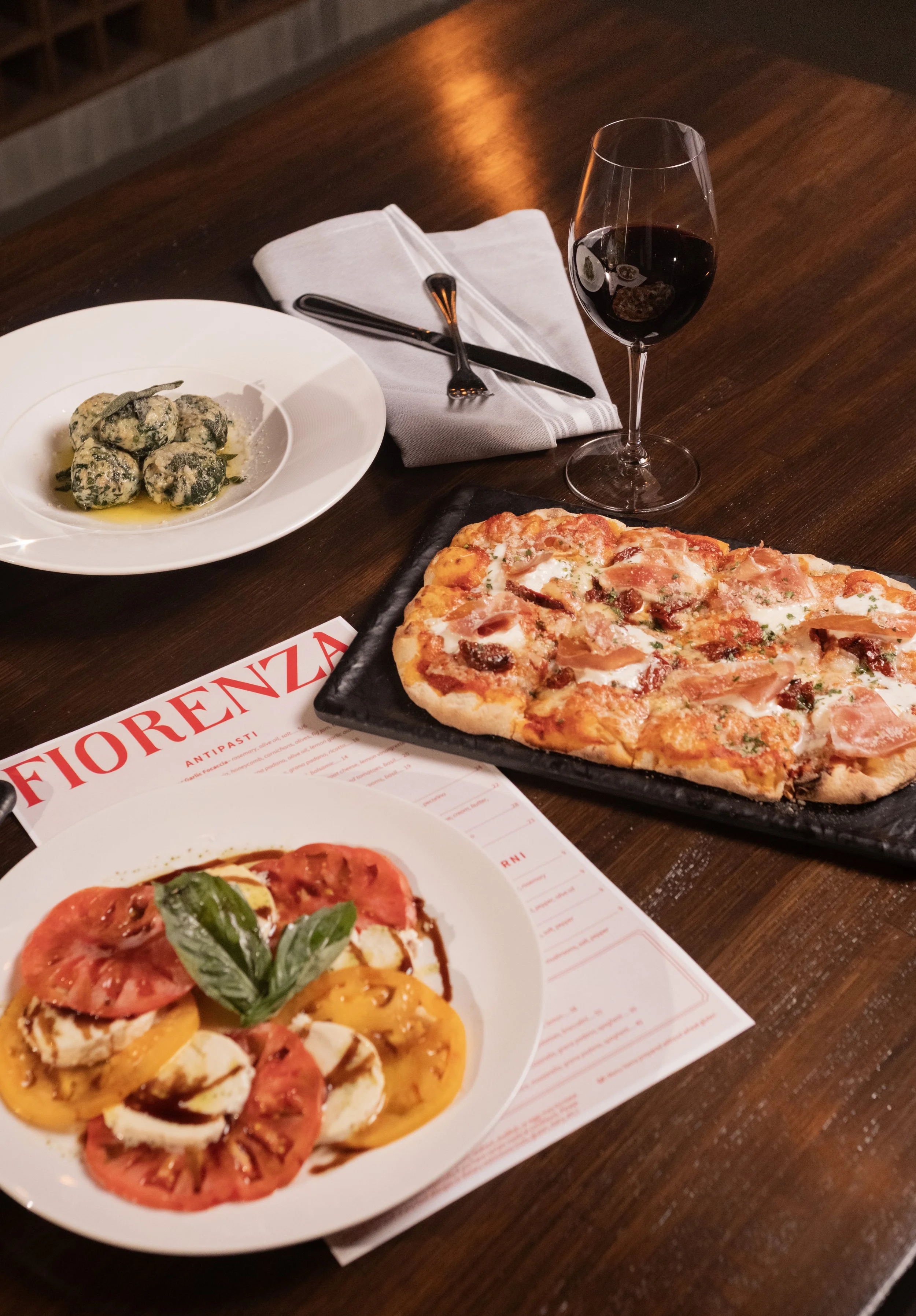 A pizza with ham, tomatoes, and basil, a plate of ravioli with spinach and cheese, a glass of red wine, a fork and napkin on a wooden table, and a menu from Fiorenzo.