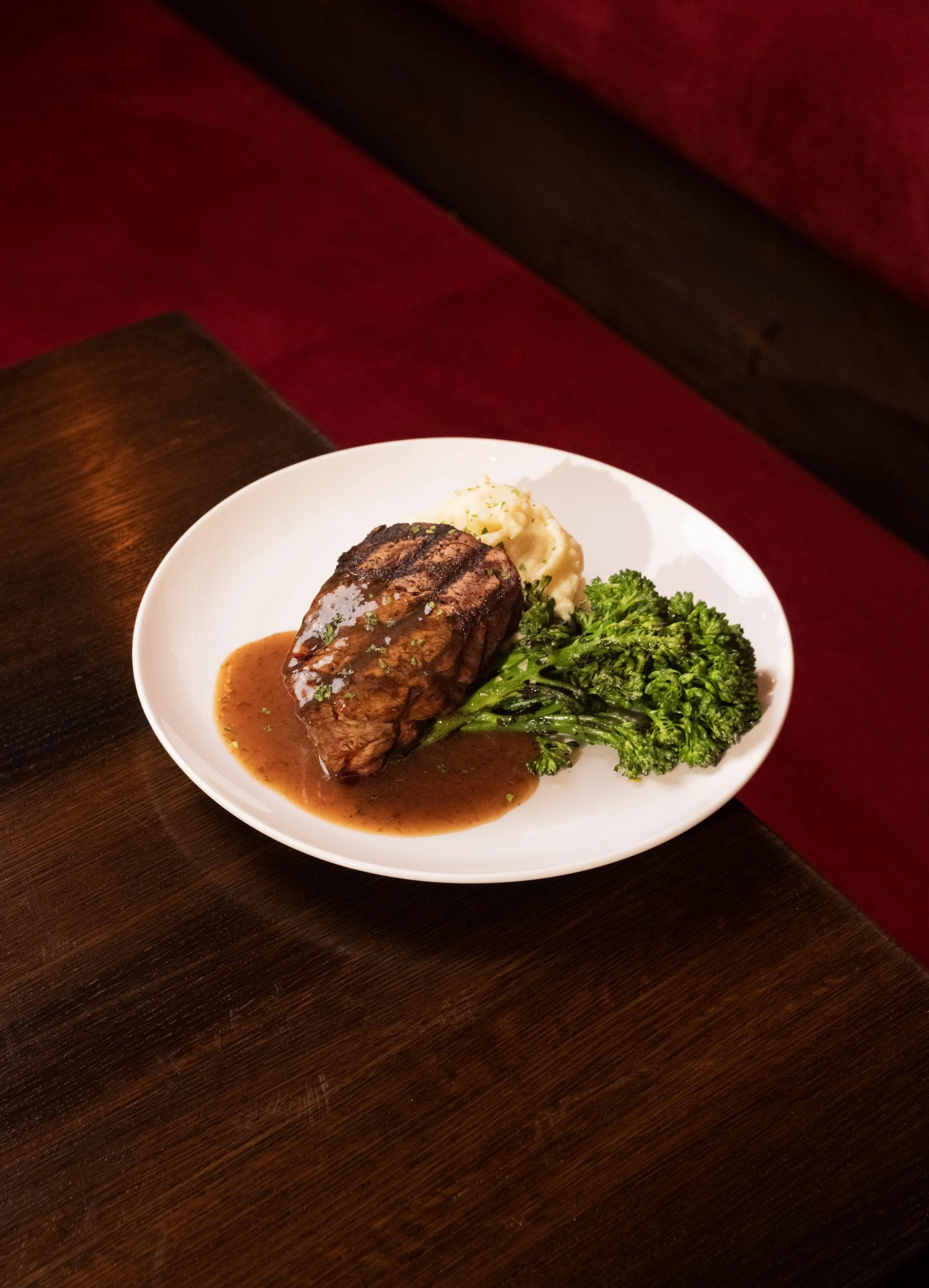 A plate of food with a piece of beef steak, mashed potatoes, and broccoli, served on a white dish on a dark wooden table with red upholstery in the background.