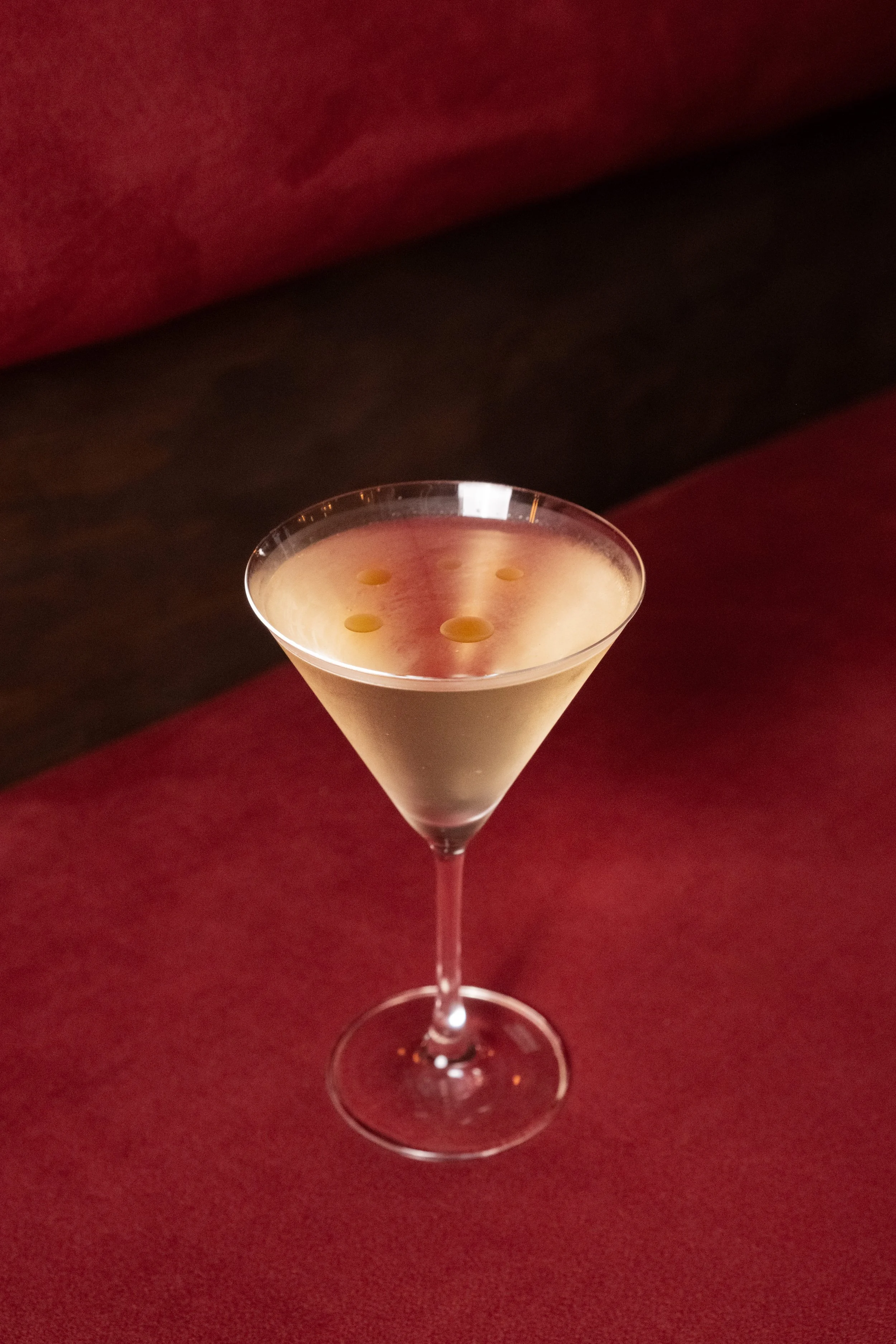 A martini glass with a light-colored cocktail garnished with three green olives, placed on a dark red surface with a dark background.