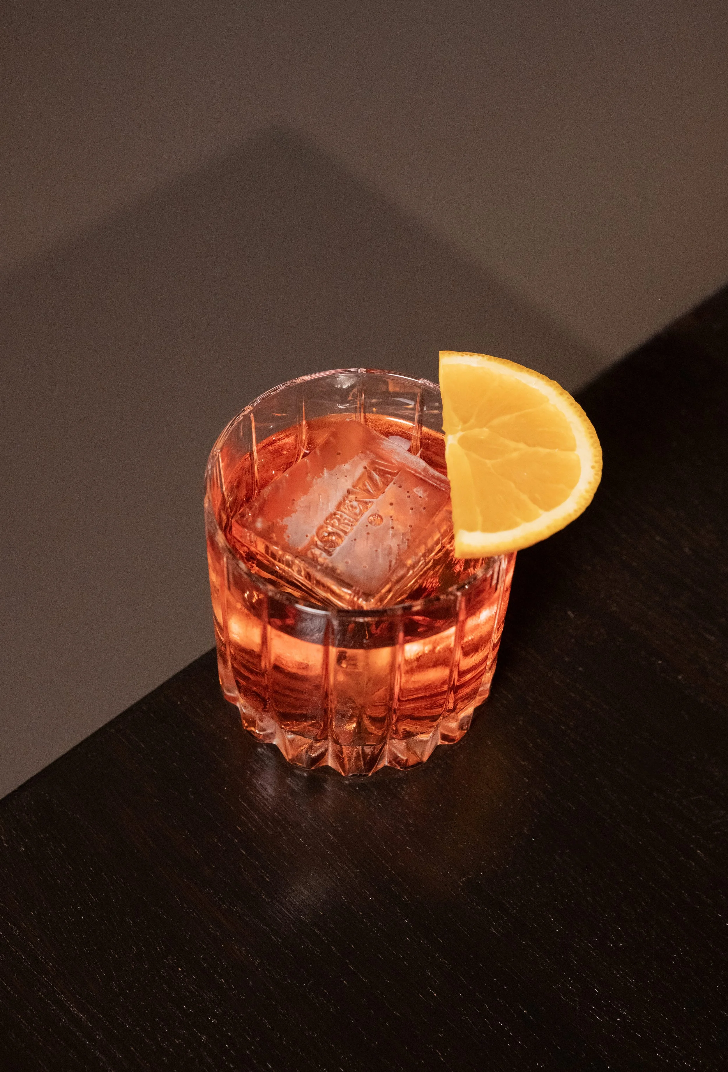 A pink-colored drink in a faceted glass with a large ice cube and an orange wedge garnish placed on the rim, sitting on a dark wooden surface.