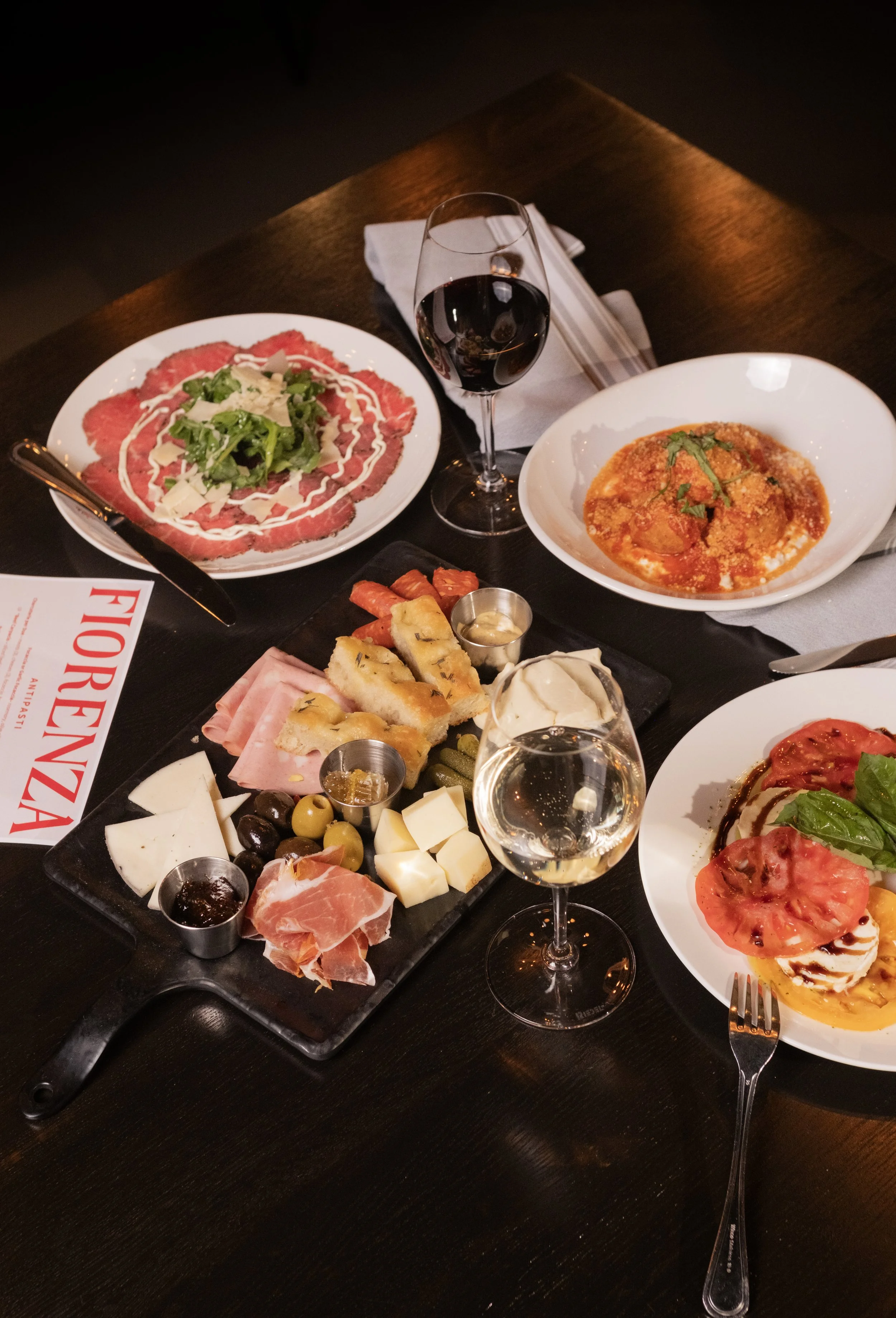 A table of Italian food including a charcuterie board with cheeses, meats, and olives, a plate of sliced beef carpaccio with greens and cheese, a bowl of ravioli in tomato sauce garnished with basil, a glass of red wine, and a small plate of sliced tomatoes topped with mozzarella and basil.