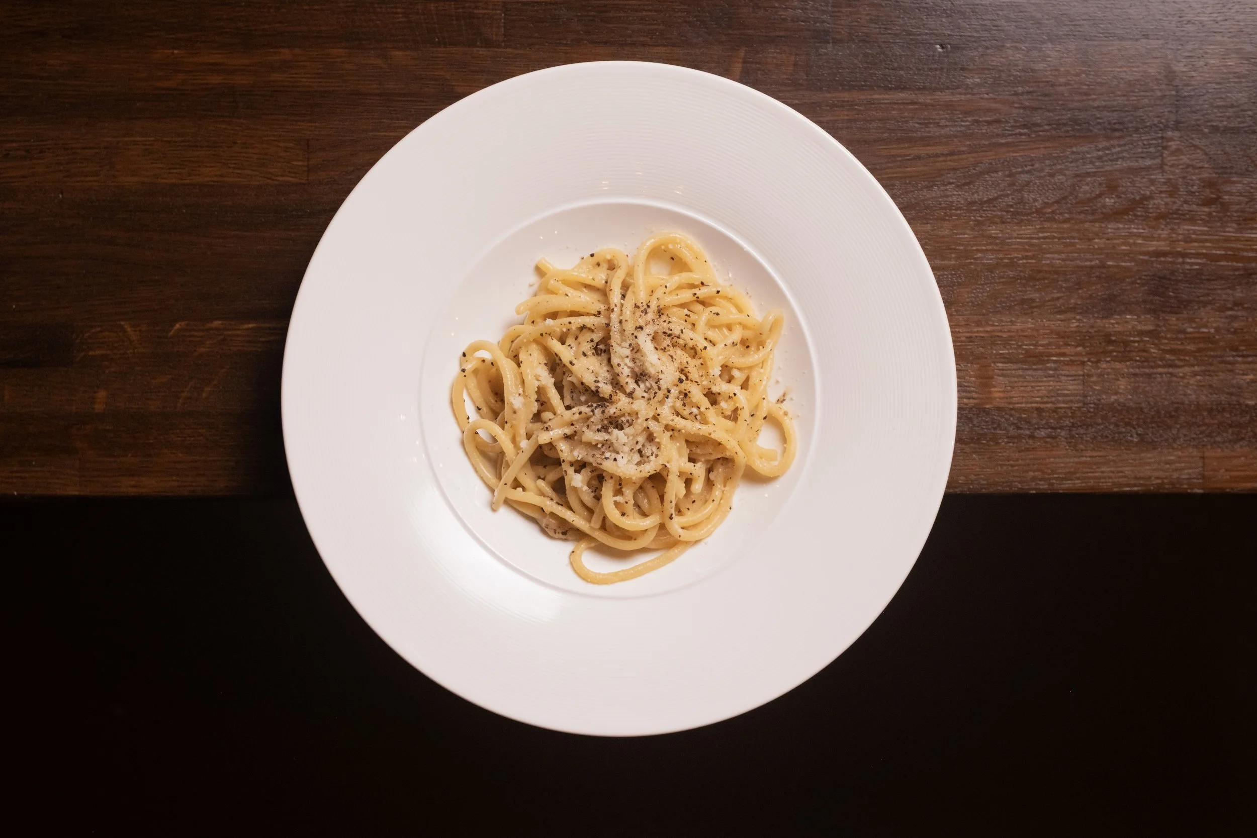 A plate of spaghetti pasta with black pepper on top, on a dark wooden table.