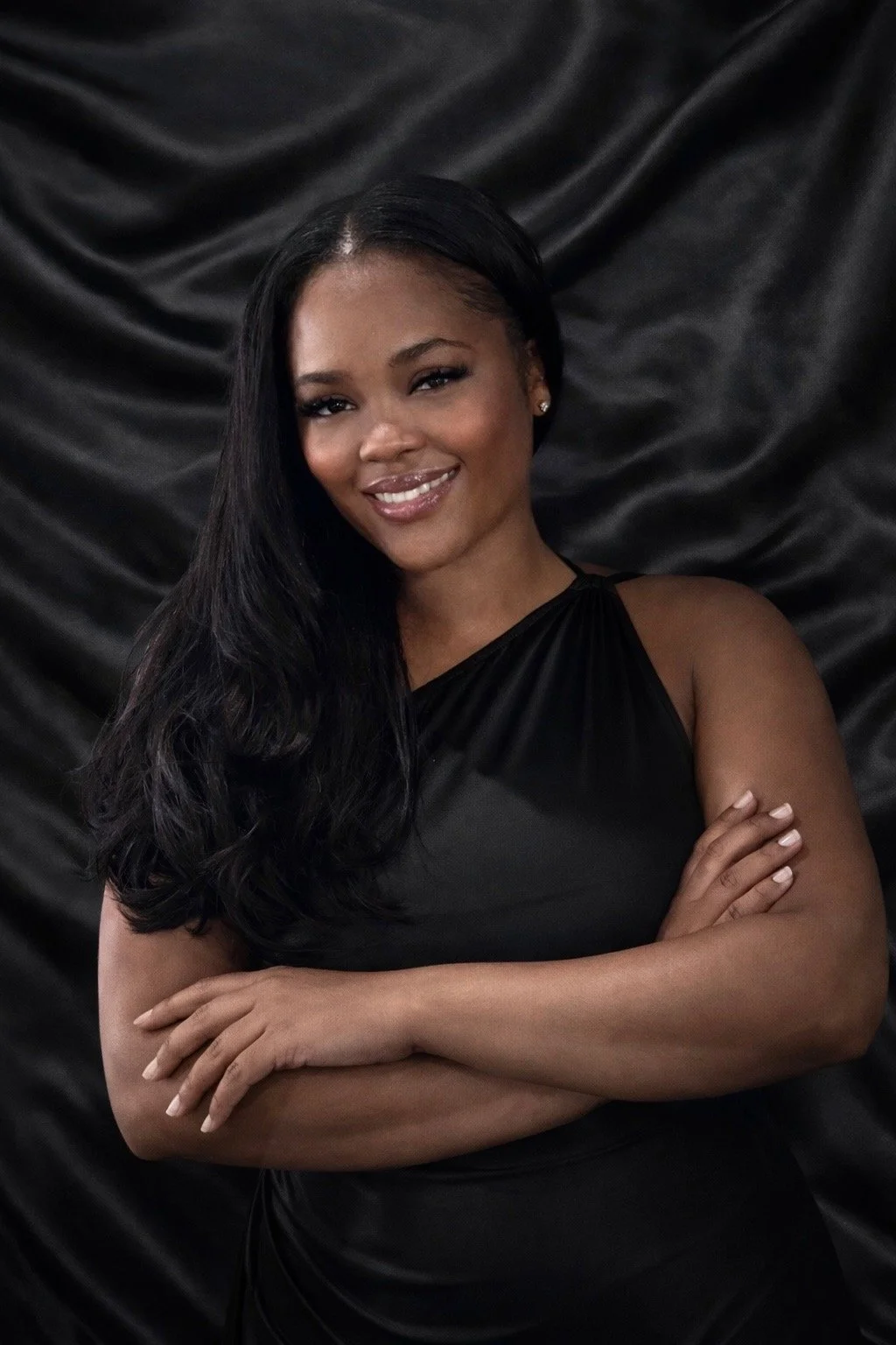 A woman with long black hair wearing a black dress, standing with her arms crossed, smiling, against a black satin backdrop.