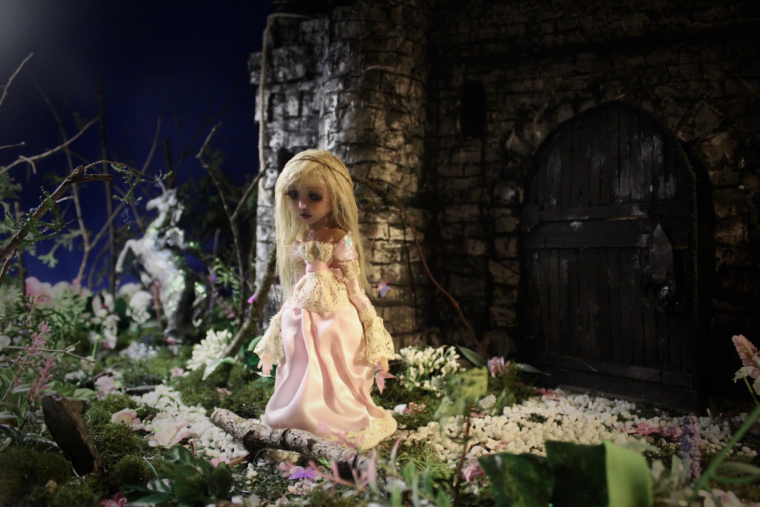 A doll in a pink dress stands among grass, flowers, and small branches near a dark stone building with an arched wooden door at night.