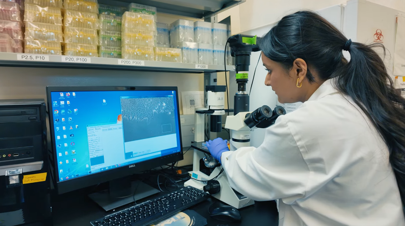 A scientist in a white lab coat examining a sample through a microscope in a laboratory. A computer monitor displays scientific data and images, with shelves of laboratory supplies in the background.