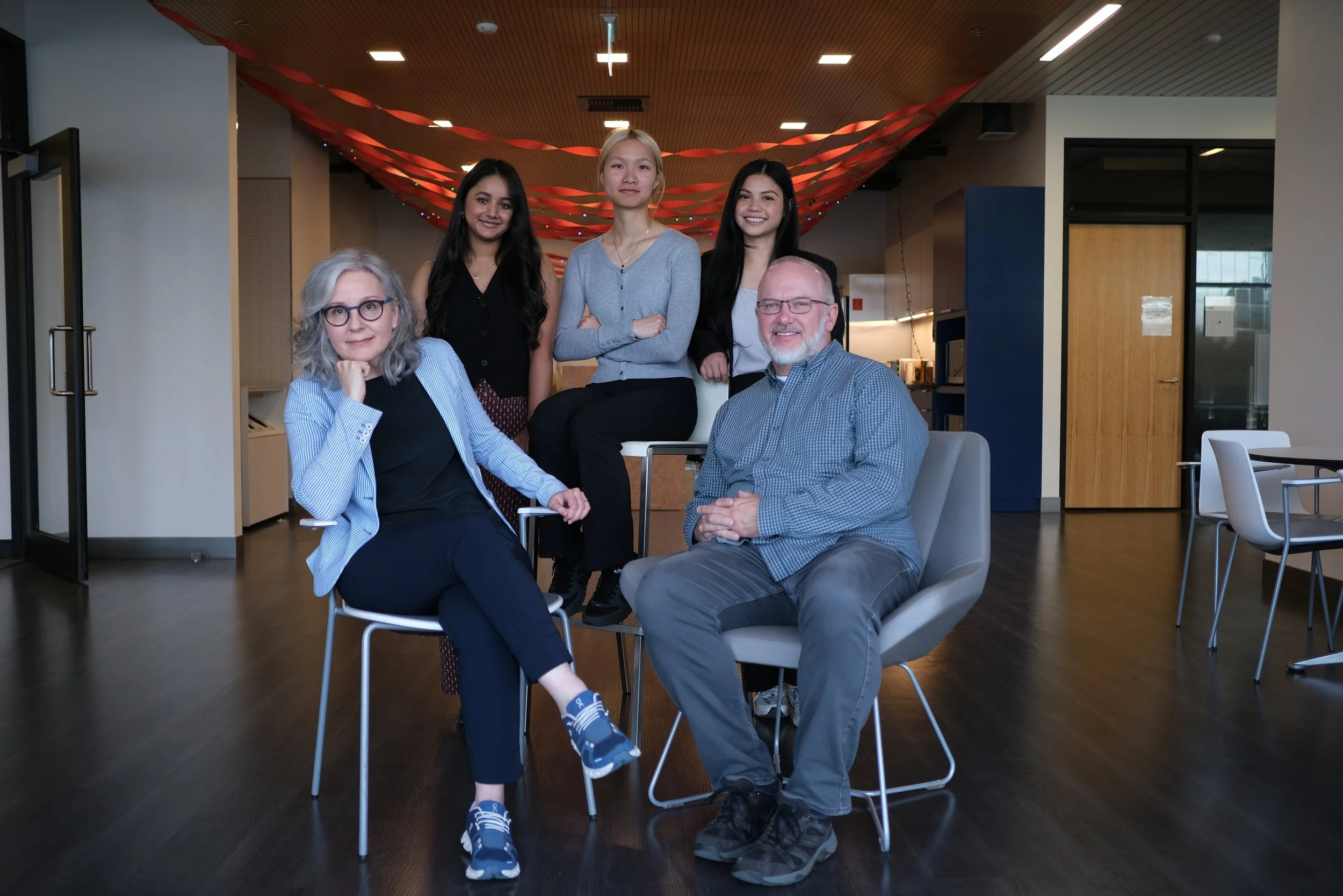 Group of five diverse professionals in an office, with three women standing behind one seated men on the right and another seated women on the left, all smiling at the camera.