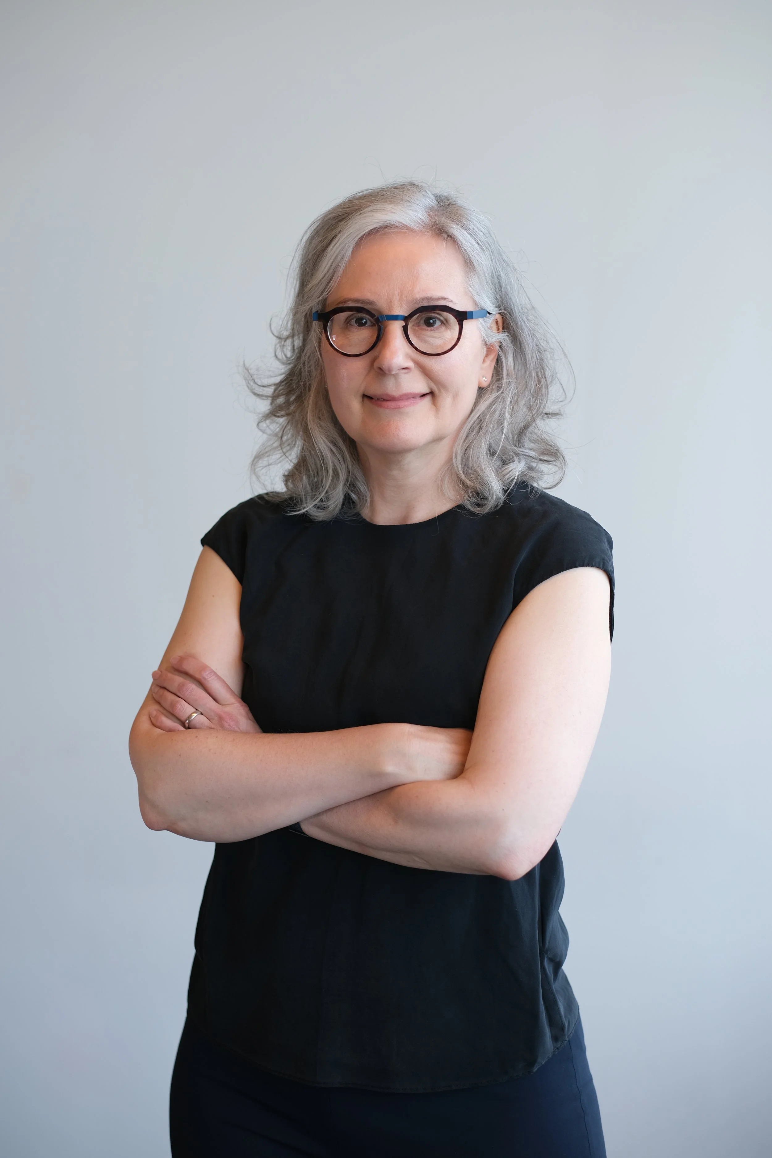 A portrait of a middle-aged woman with gray hair and glasses, wearing a black top, standing with arms crossed against a plain light-colored background.