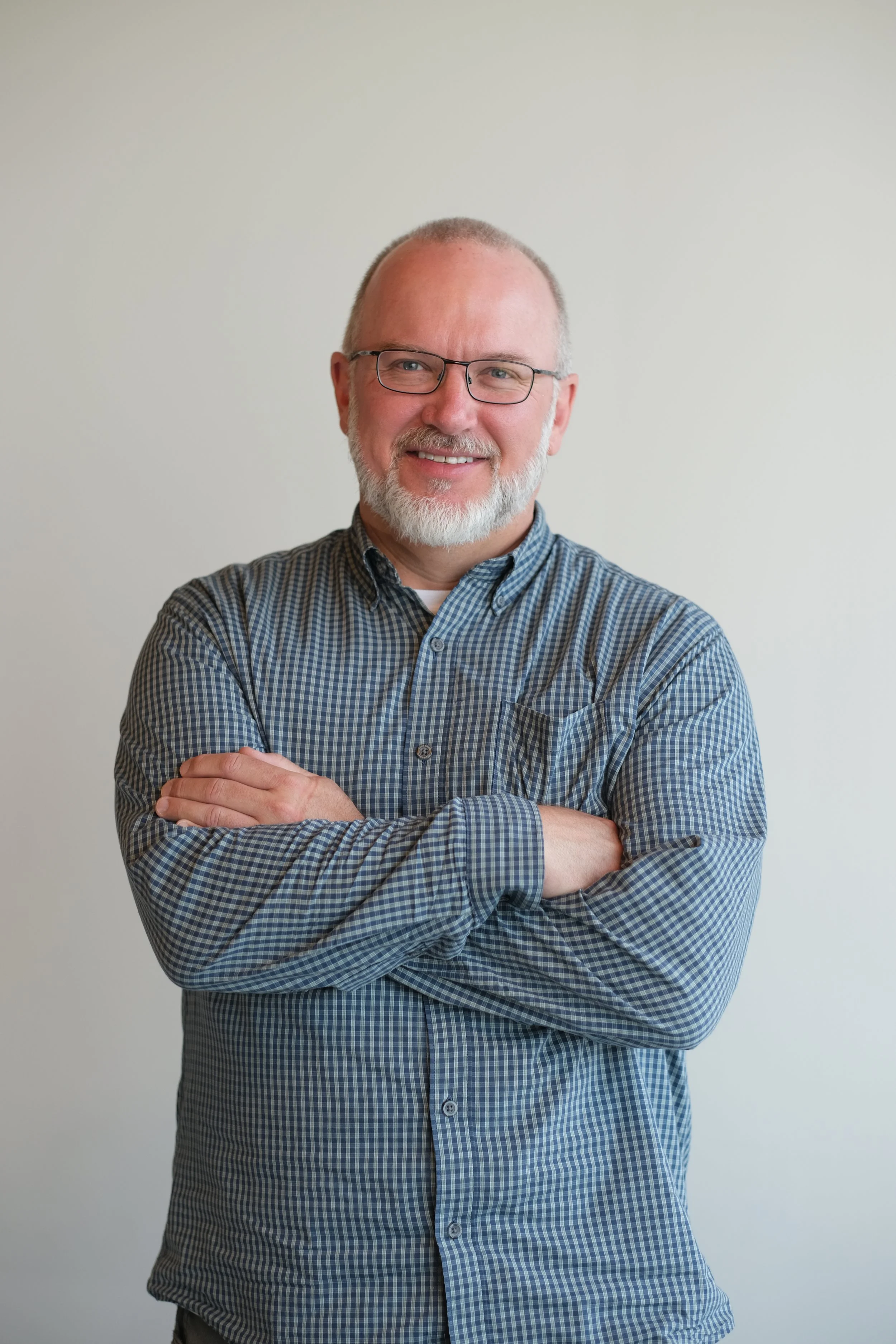 A middle-aged man with glasses, a beard, and short hair, smiling and crossing his arms, wearing a checkered shirt, standing against a plain light-colored background.