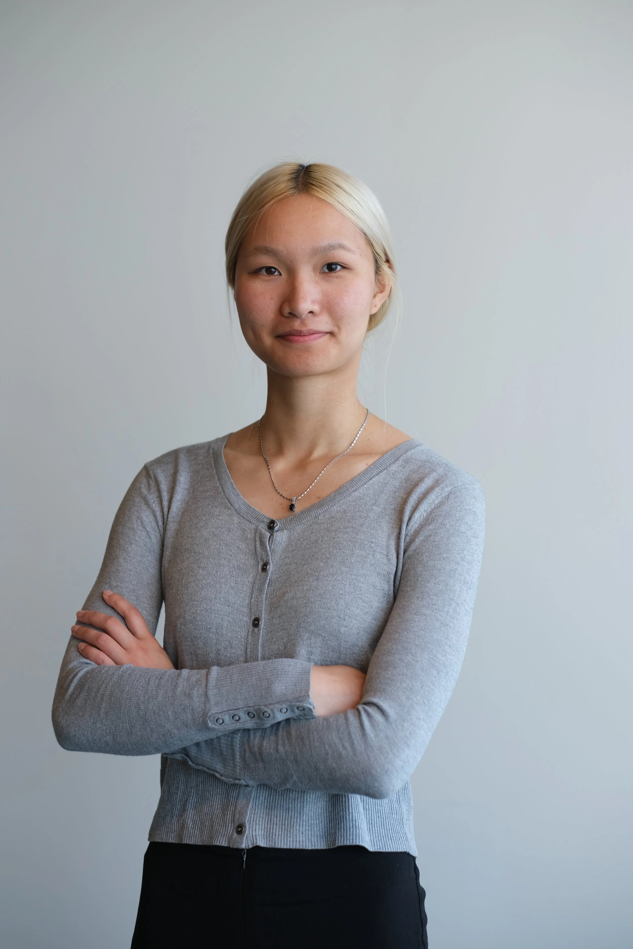 A young woman with blonde hair standing with arms crossed, wearing a gray cardigan and black pants, against a plain light-colored background.