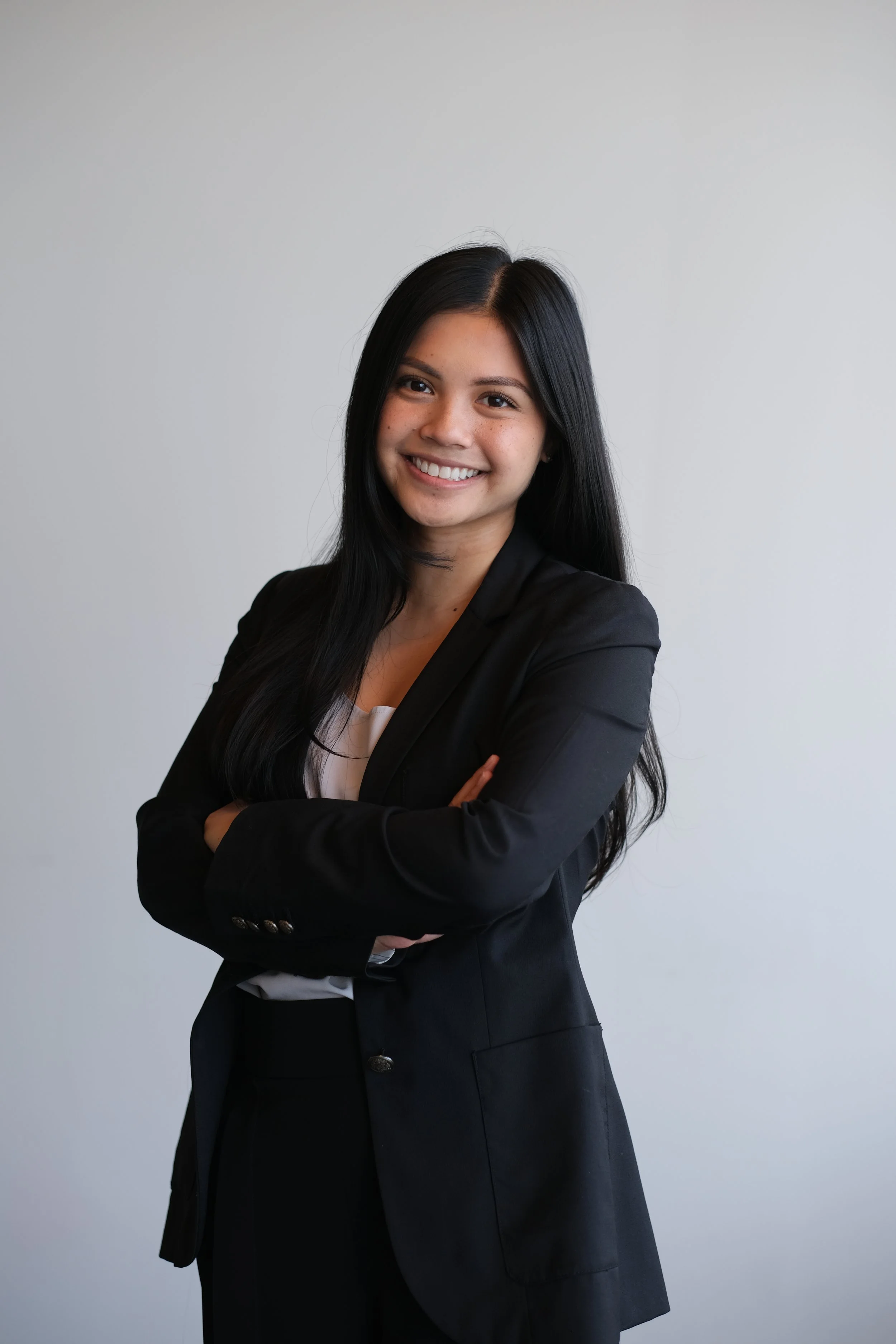 A young woman with long black hair, smiling, in a black blazer and white shirt, standing against a plain light gray background, with arms crossed.