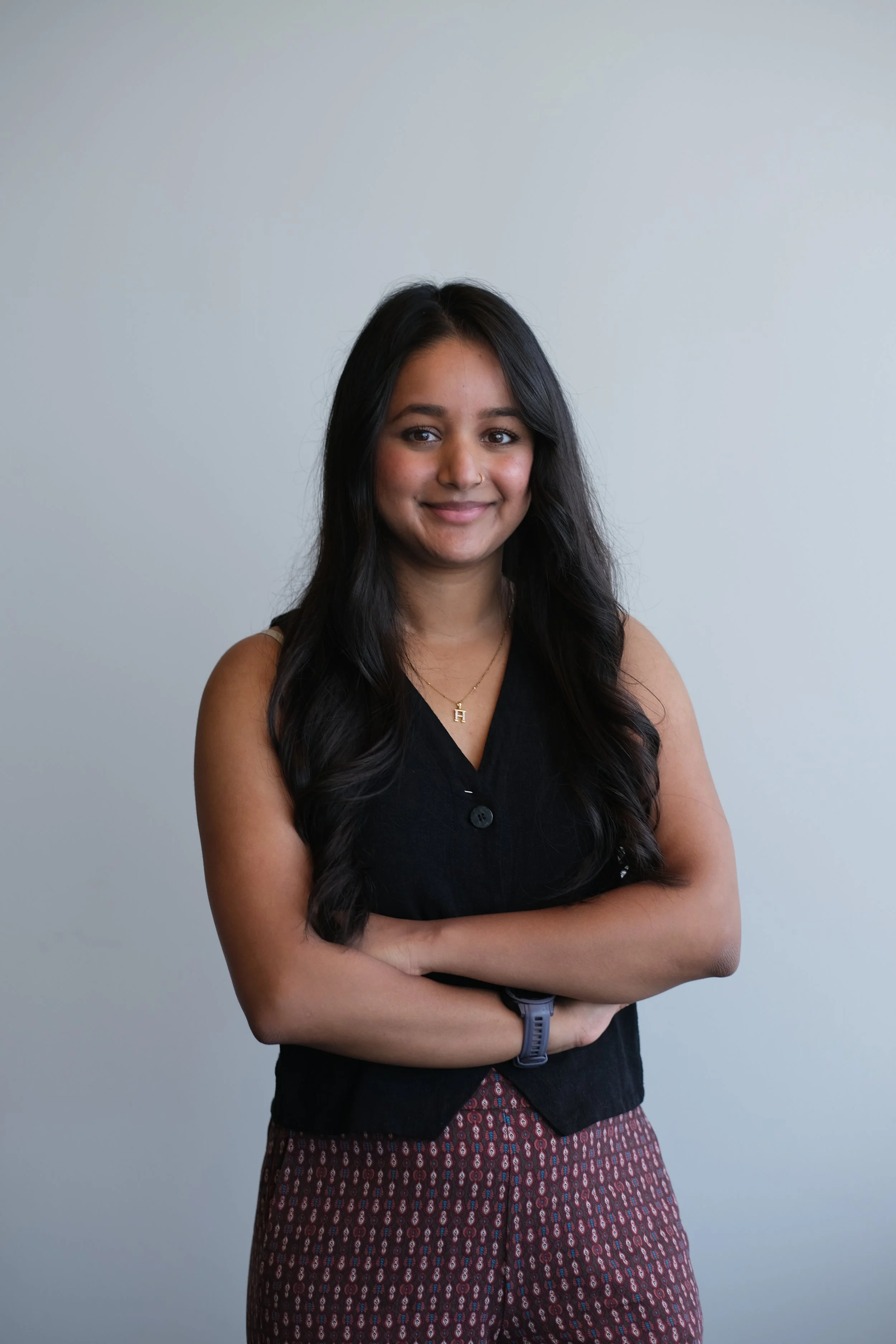A woman with long dark hair smiling, wearing a black sleeveless top with a necklace, patterned pants, and a wristwatch, standing with arms crossed against a plain light gray background.