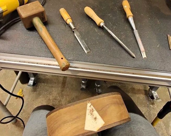 A workbench with four woodworking tools: a mallet and three chisels with wooden handles. Below the bench is a wooden chair with a piece of wood resting on the seat.