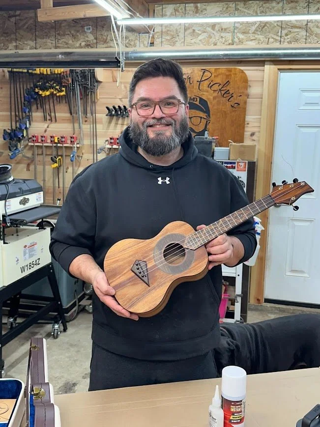 A man with glasses and a beard holding a small wooden ukulele in a workshop with tools on the wall behind him.