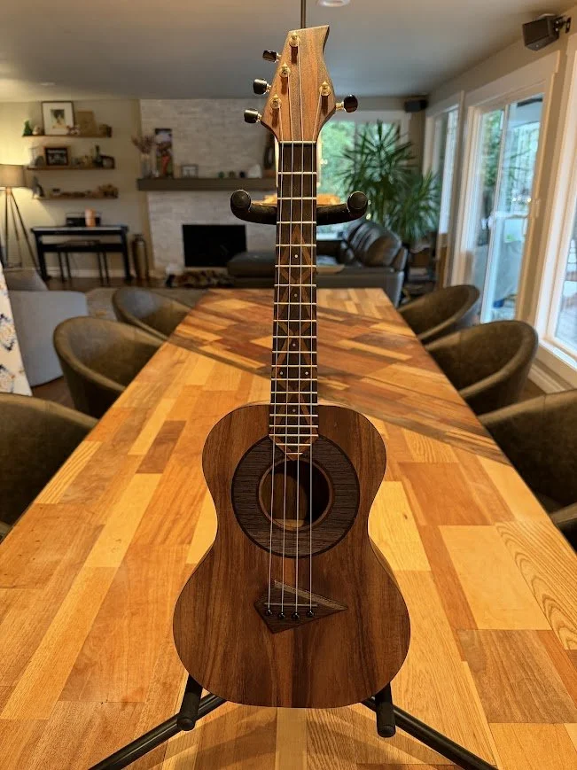 A small wooden ukulele stands on a table in a well-lit dining room with chairs around it, a stone fireplace, and large windows in the background.