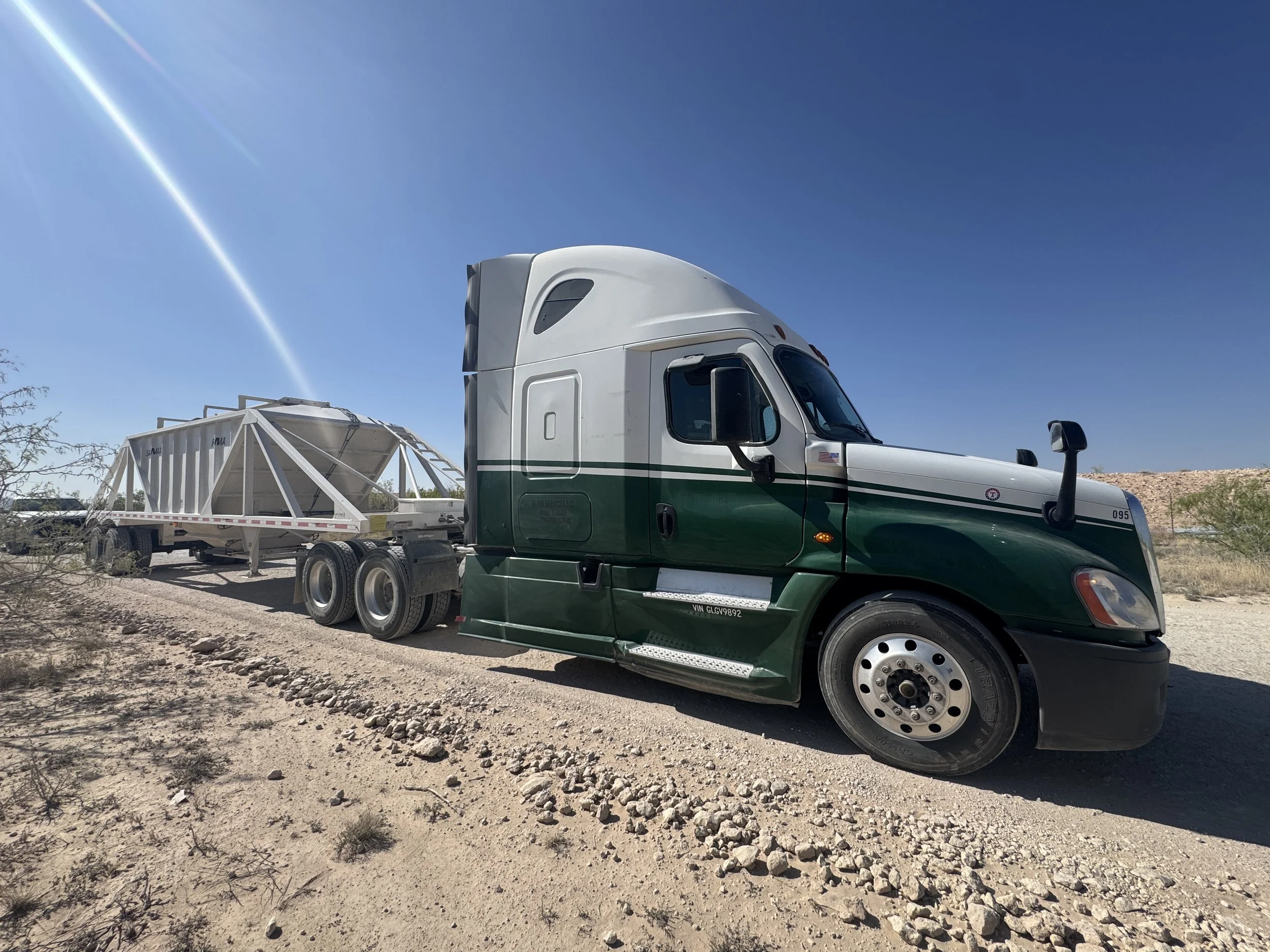 Semi-truck with a green and white cab parked on a dusty, rocky road in a desert landscape, with a trailer carrying a large metallic frame structure and a clear blue sky overhead.