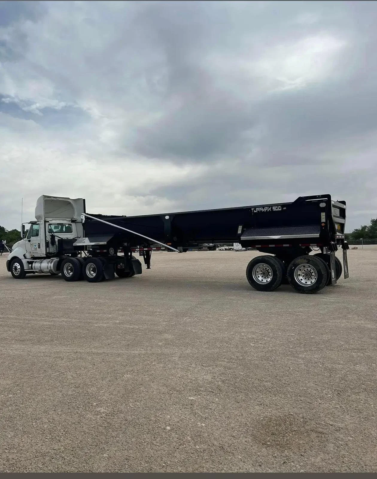 A white semi-truck with a black trailer parked on a gravel lot under cloudy skies.