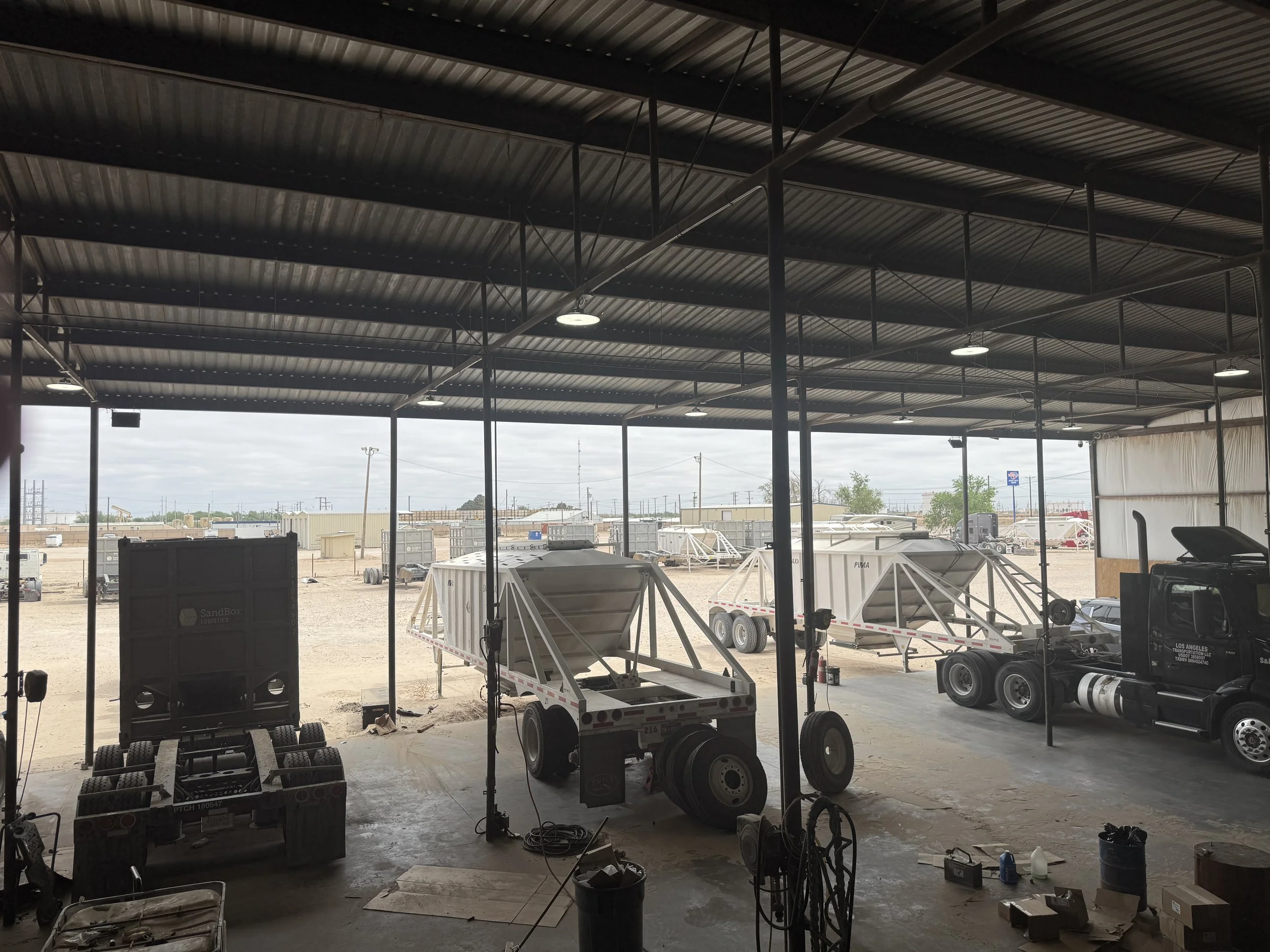 Inside a large covered industrial warehouse with a metal roof, multiple trailers and trucks are parked outside on a dirt lot, some with equipment or cargo containers, with electrical infrastructure and equipment in the background.