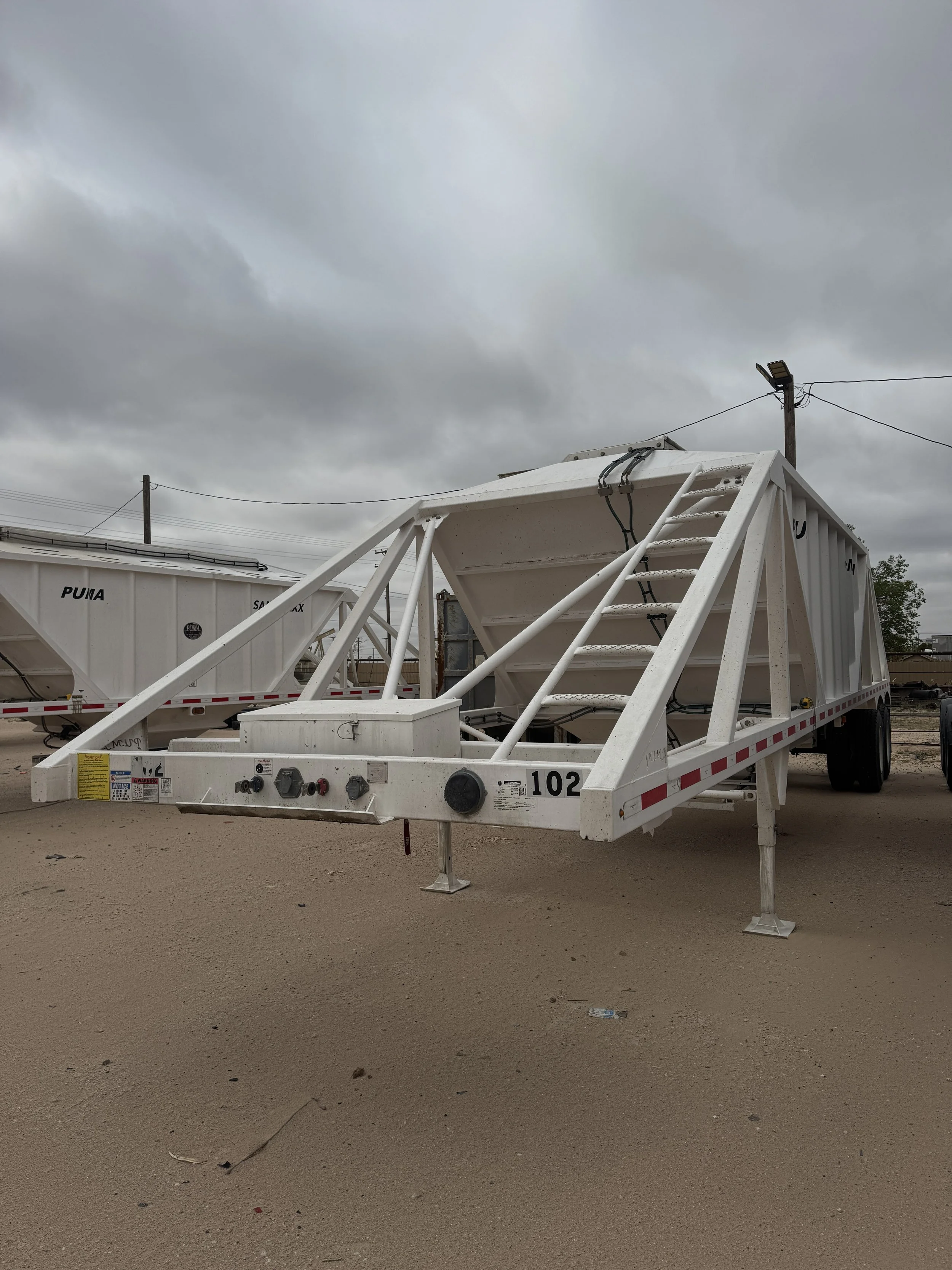 A white industrial dump trailer parked on a dirt lot, with overcast skies overhead.