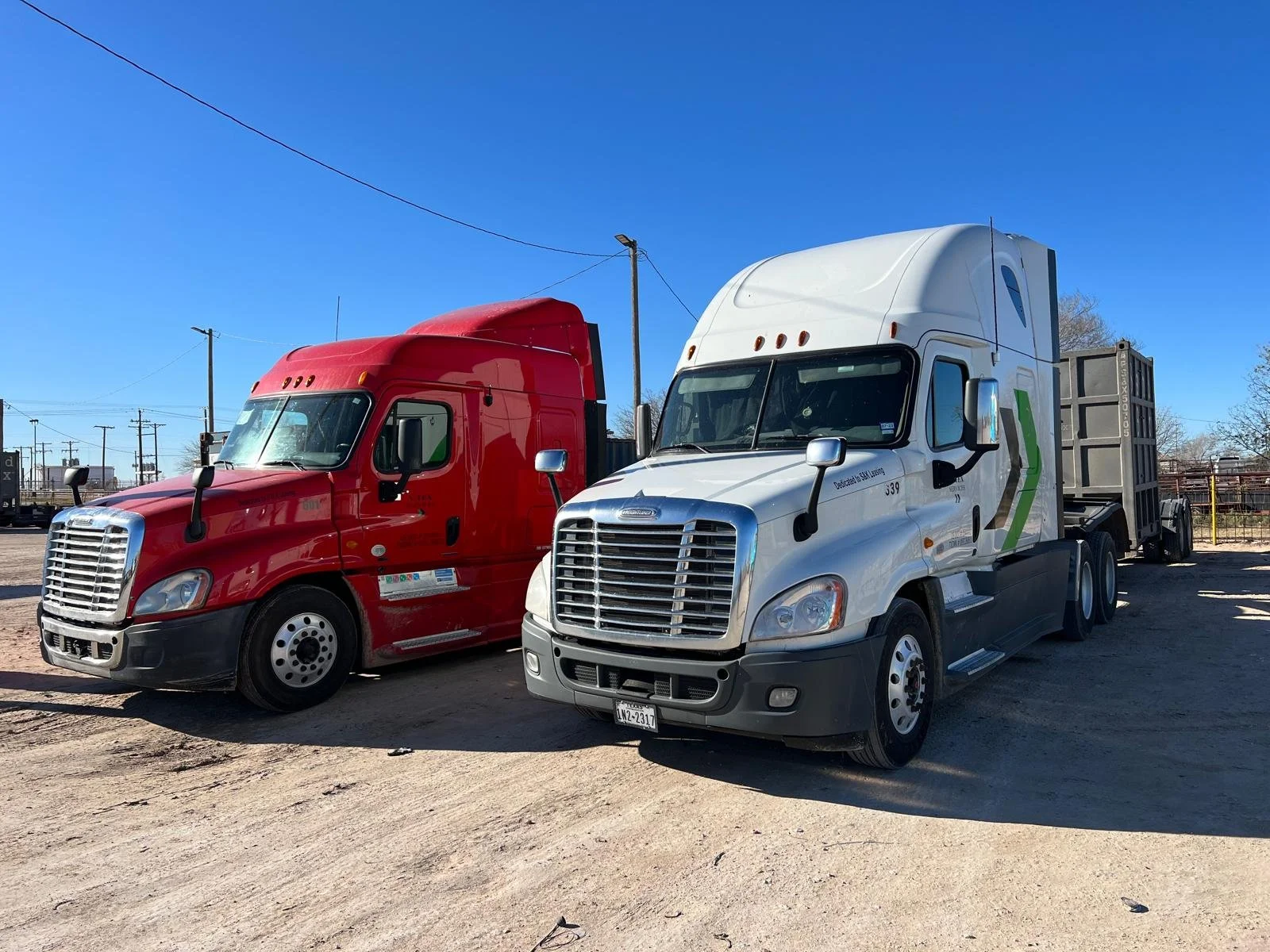 Two semi-trucks parked side by side on a dirt lot against a clear blue sky, one red and one white.