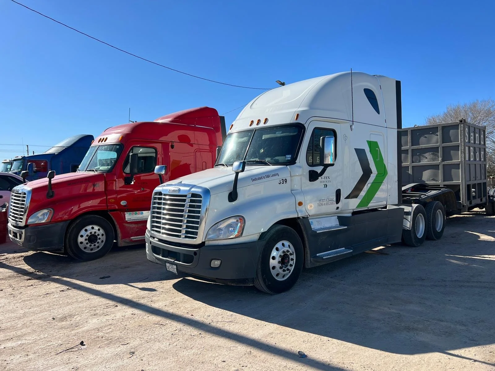 A row of semi-trucks parked on a dirt lot under a clear blue sky, with a white truck in the foreground and red and blue trucks behind it.