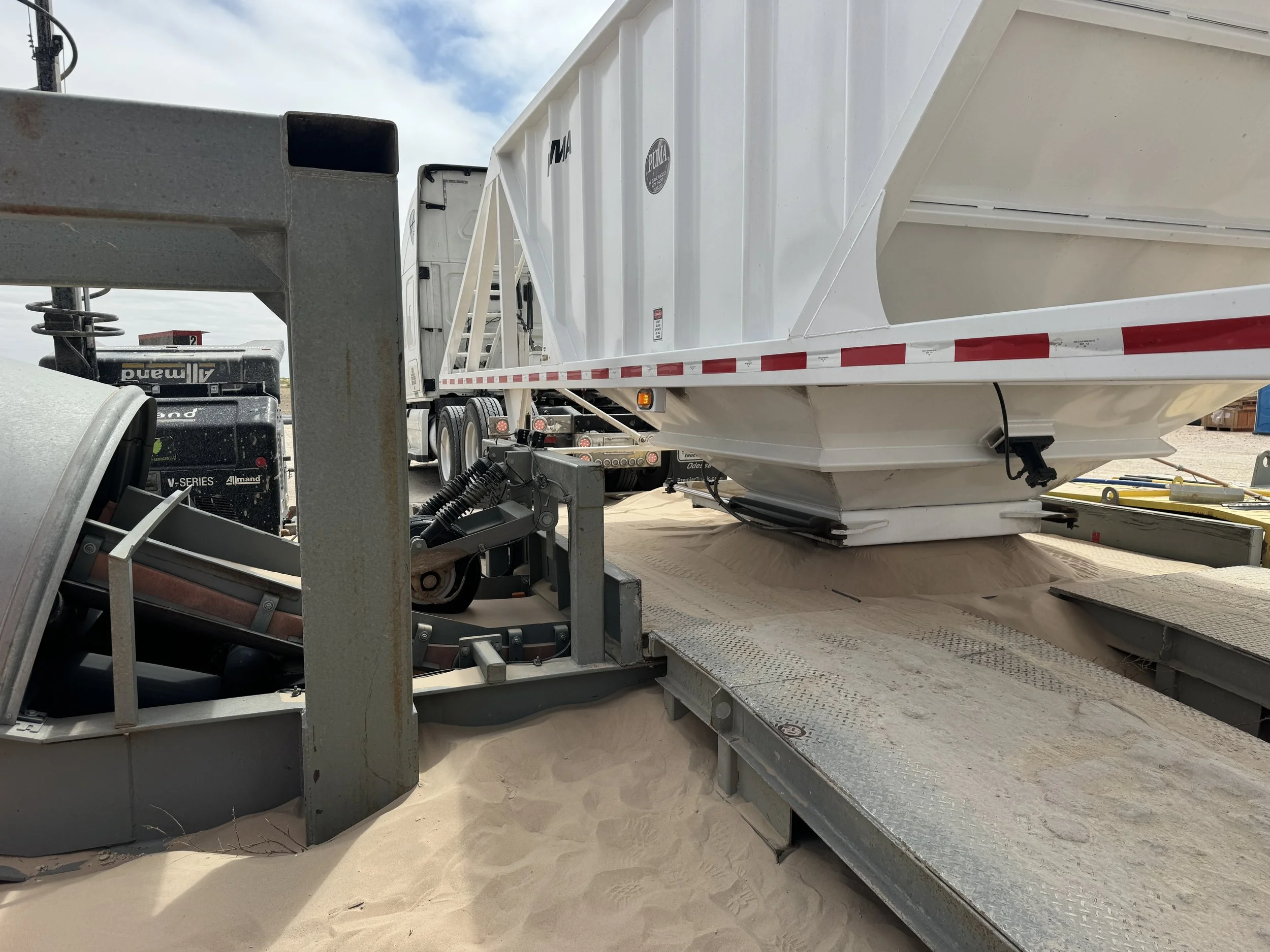 Close-up of a boat trailer and boat on a sandy surface, with trucks and equipment in the background.