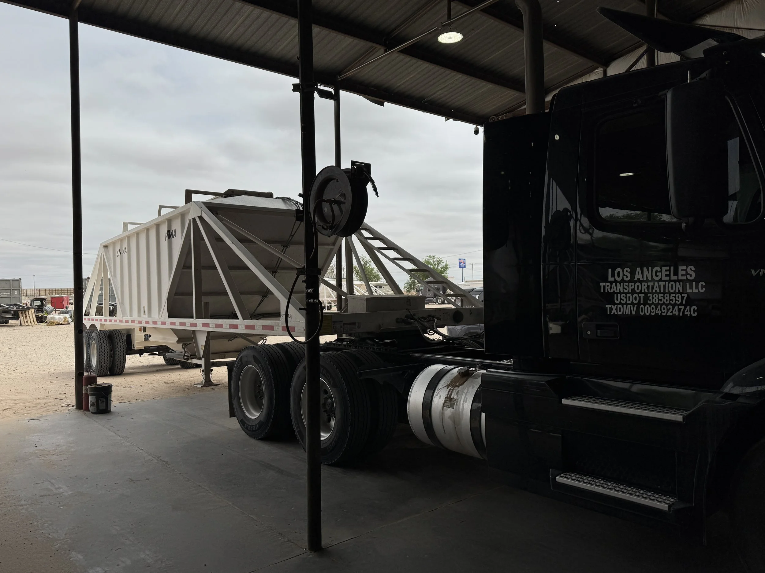 A black semi-truck with a white trailer parked inside a covered area, with supplies and equipment visible in the background outside.
