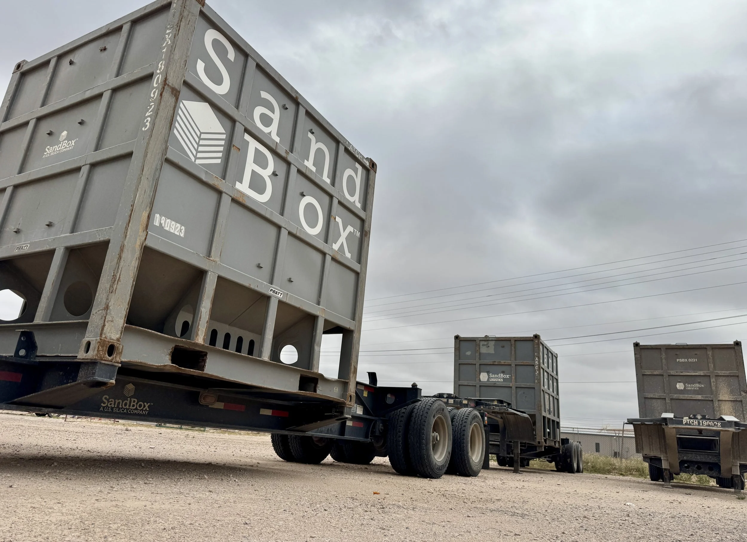 A semi-truck with an empty flatbed trailer parked on a gravel lot under a cloudy sky, with large Boddie Load Container boxes on the trailer and nearby.