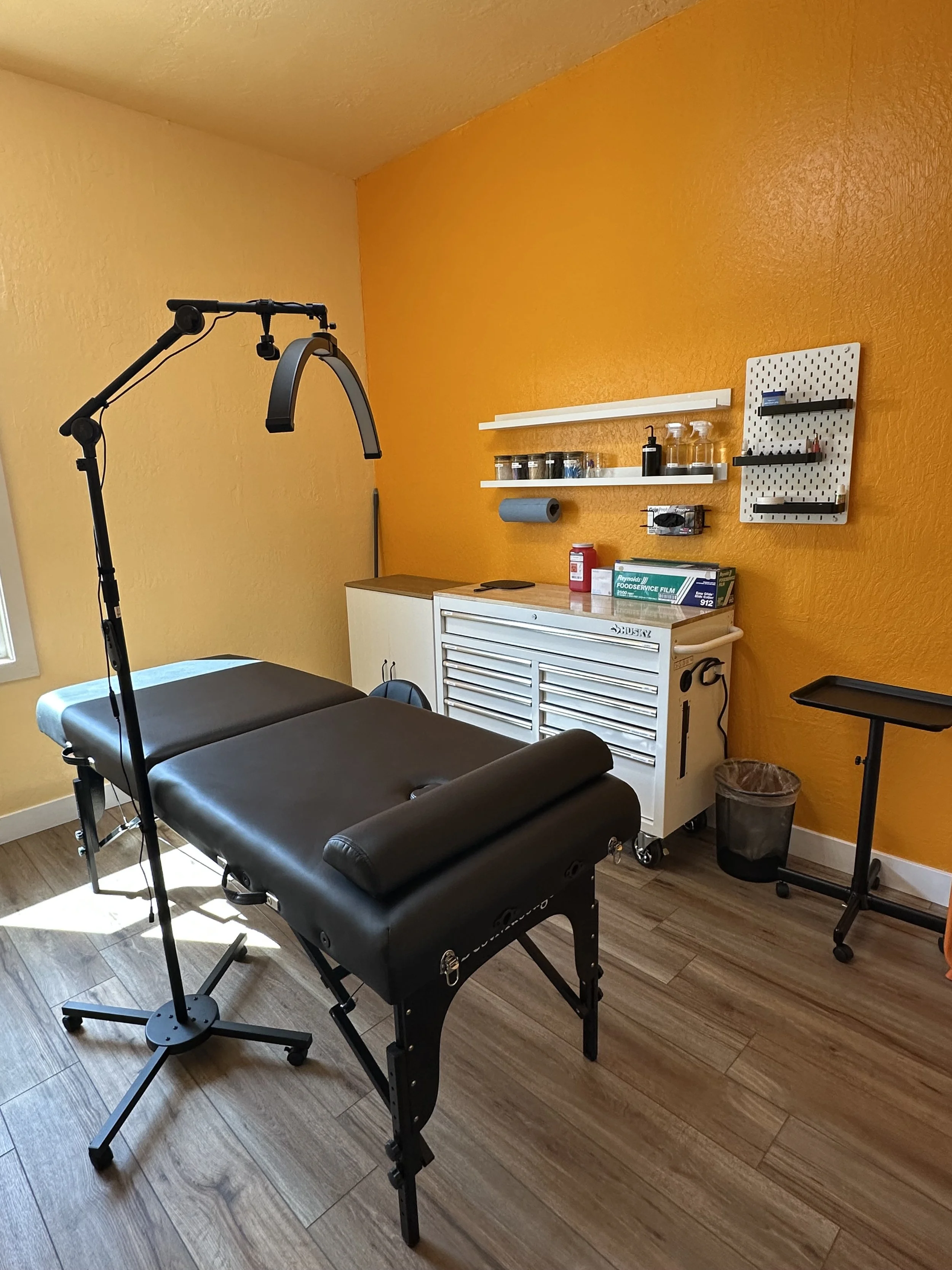 A treatment room with a massage table, black, in front of yellow and orange walls. There is a small rolling tray, shelves, a cabinet, and various small containers and supplies on the wall and counter.
