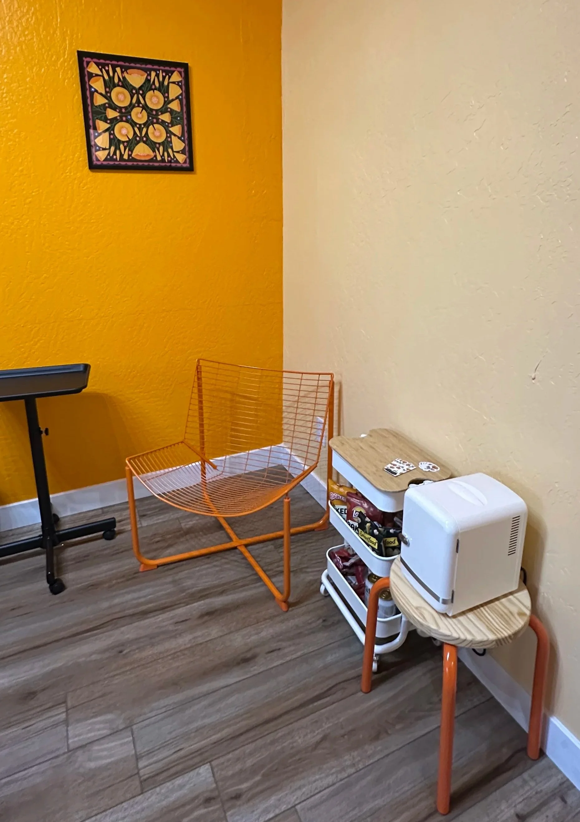 A corner of a room with yellow and beige walls, a small orange wire chair, a wooden side table with a white wine cooler, and a shelving unit with snacks and drinks. There is a colorful abstract painting on the yellow wall.