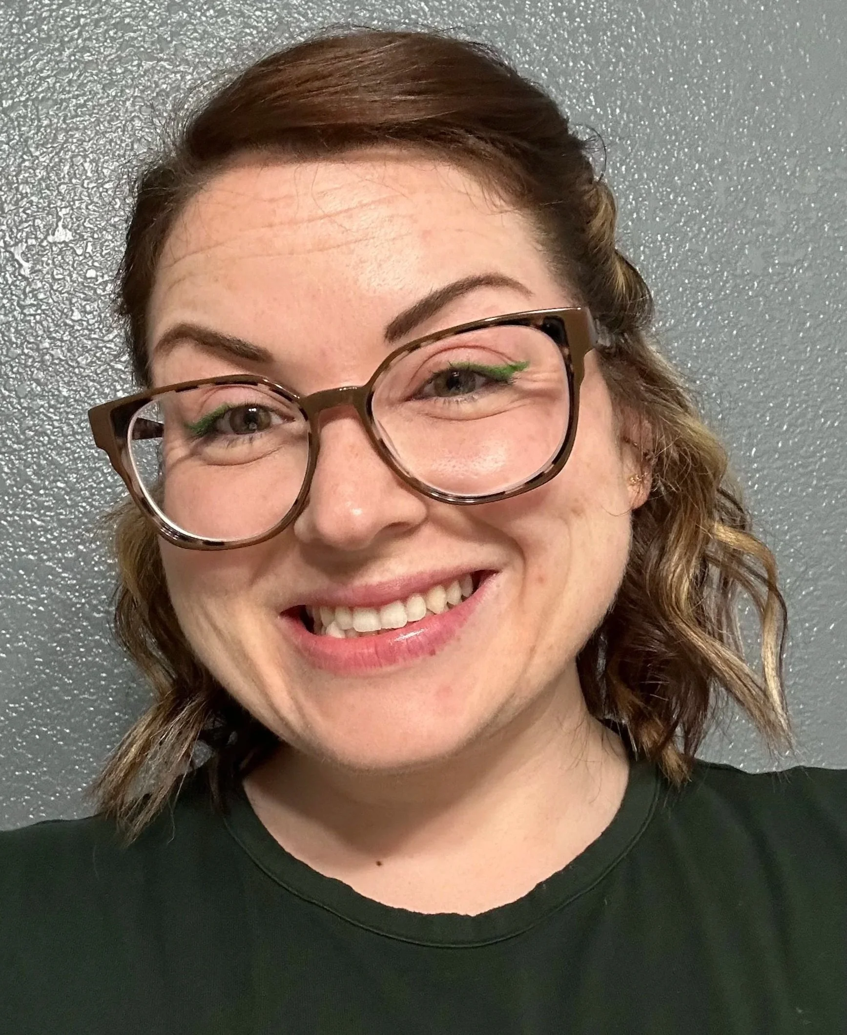 Close-up of a woman with brown hair, glasses, and green makeup, smiling in front of a gray textured wall.