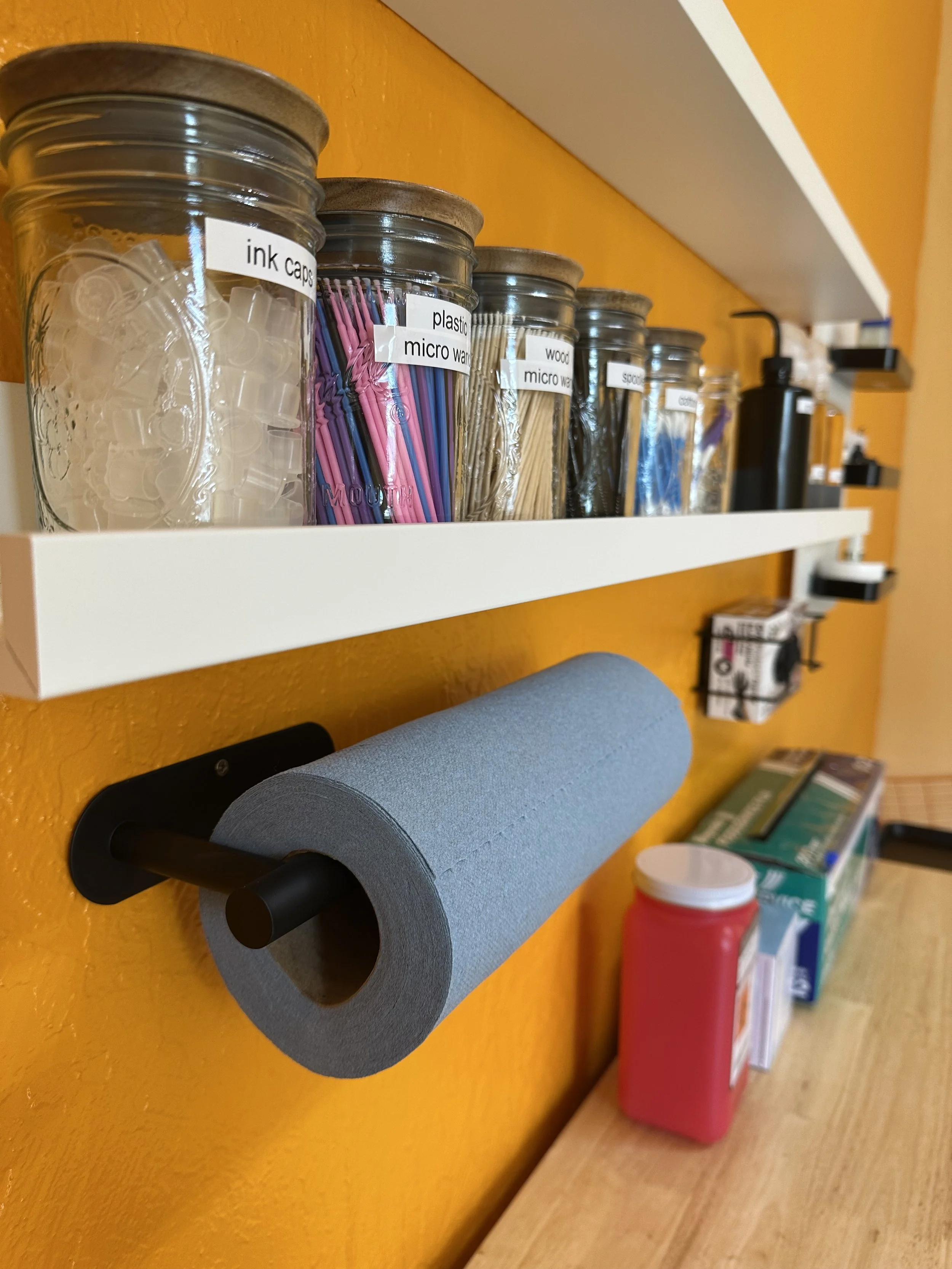 A wall-mounted paper towel holder with a roll of blue paper towels. Above it, a white shelf holds jars labeled with various contents, including ink caps, plastic micro-wand, wood micro-wand, spoon, and scraper. There are additional supplies on the counter below the shelf, such as a red bottle and boxes.