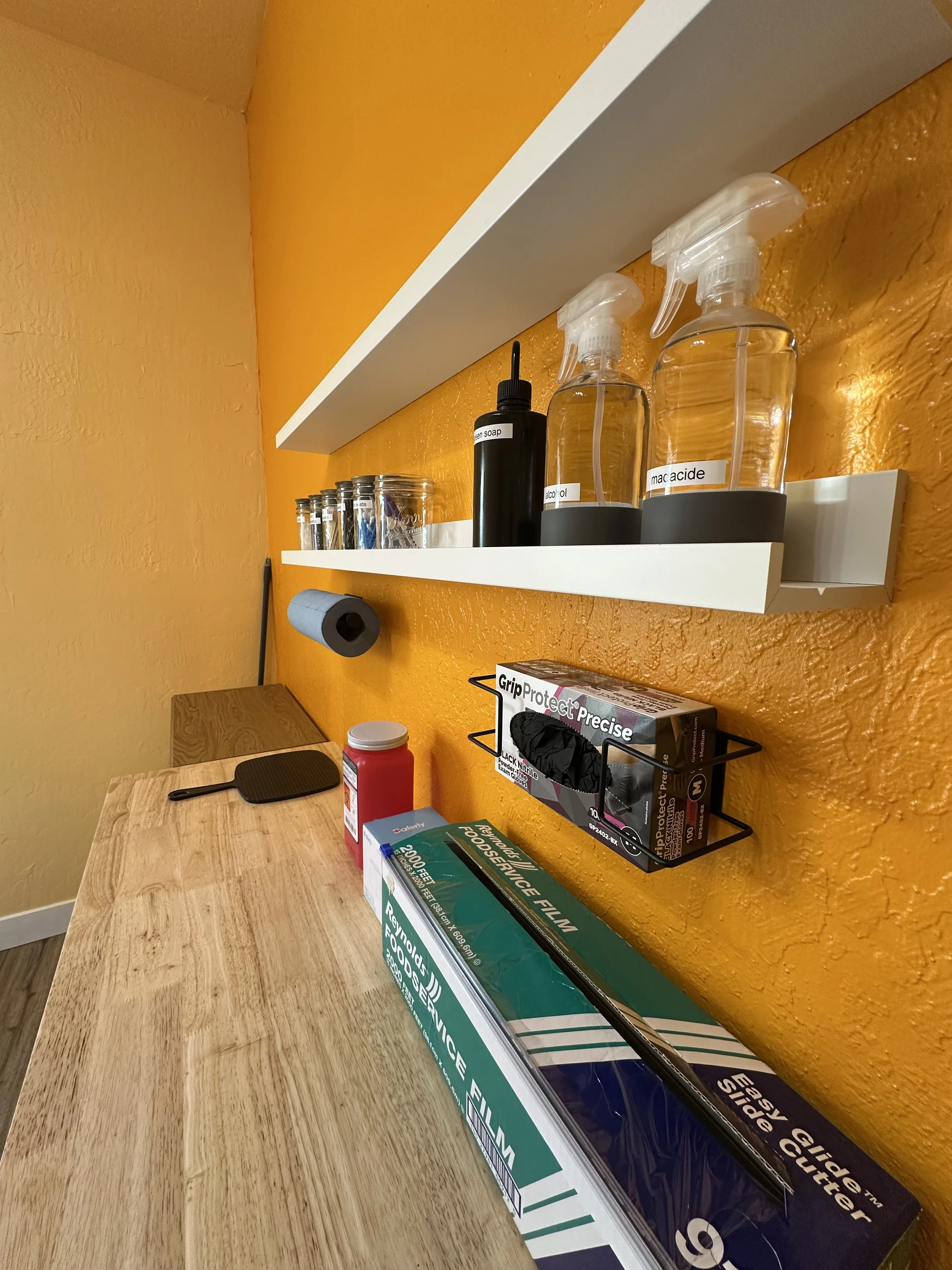 A wooden counter with various cleaning and safety supplies, including spray bottles, a roll of blue paper towels, latex gloves, and a box of plastic gloves, against an orange wall with a white shelf holding jars and small bottles.