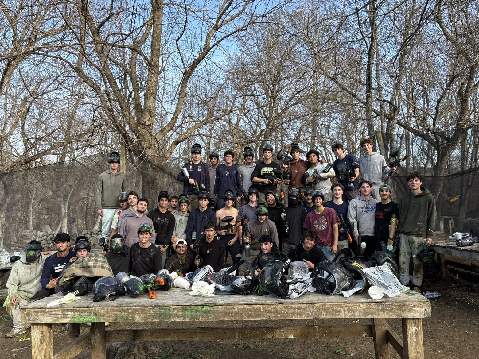 Fraternity brothers posing with paintball gear outdoors in a wooded area, some holding paintball guns, helmets, and masks, with helmets and equipment on a table in front.