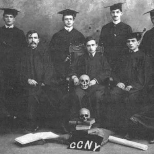 Group of fraternity founders in caps and gowns with a skull, books, and shirt labeled 'CCNY' in front.