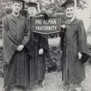 Three graduates in caps and gowns holding a Phi Alpha Fraternity sign.