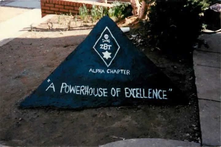 Painted triangular stone with a skull and star emblem, labeled 'ZBT Alpha Chapter,' with the quote 'A POWERHOUSE OF EXCELLENCE' written underneath.